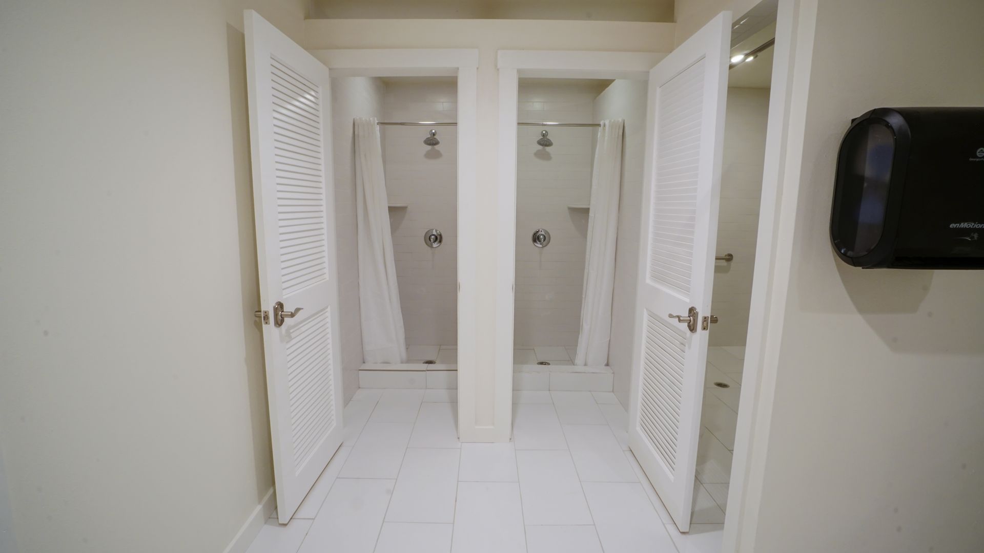 White-walled public shower room with two stalls, each with a curtain and door.  A paper towel dispenser is on the wall.