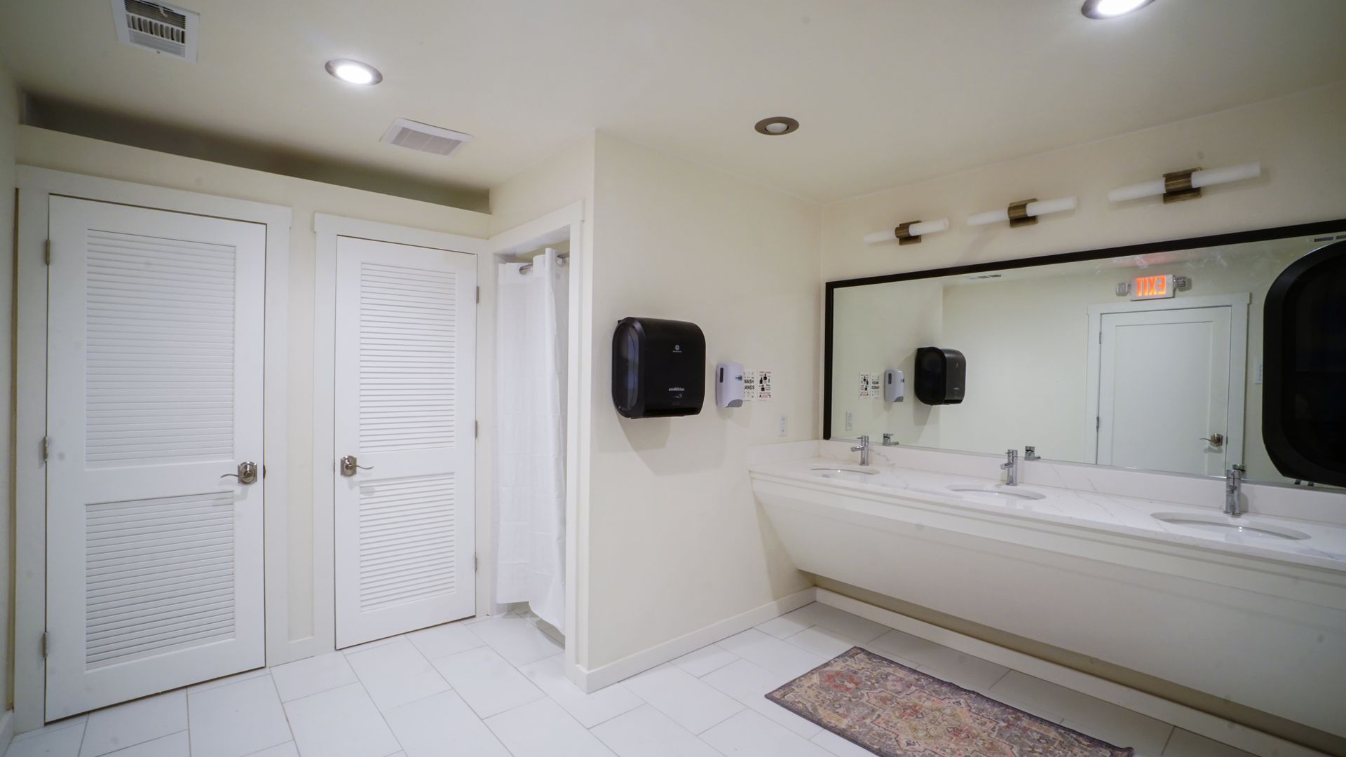 Public restroom with white walls, doors, and sinks. Black soap dispenser, mirror, and lights.