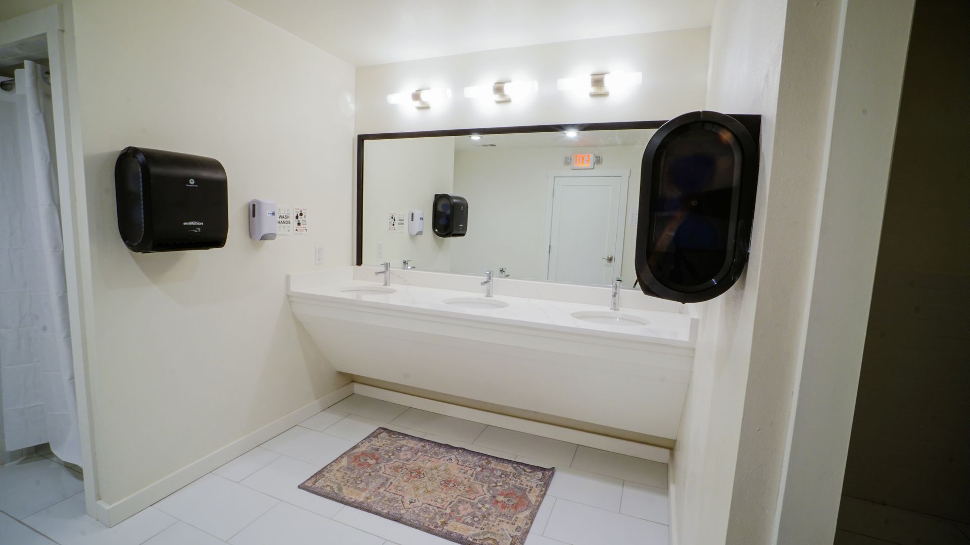 Public restroom with three sinks, large mirror, and hand dryers. White walls and tiled floor.