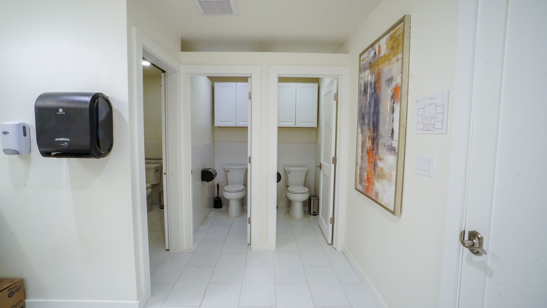 Hallway with two open toilet stalls, white walls, and a paper towel dispenser.
