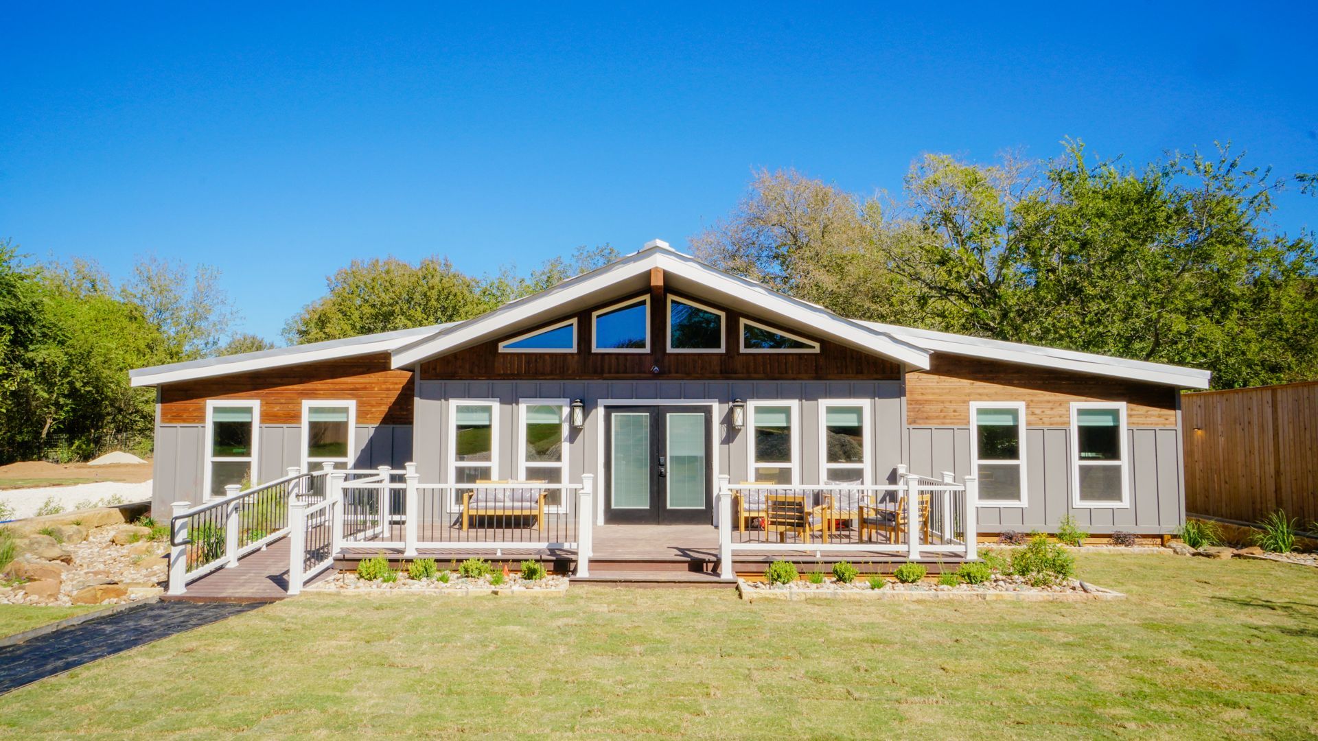 Gray house with wooden accents and a ramp leading to a porch, set on a green lawn with trees.