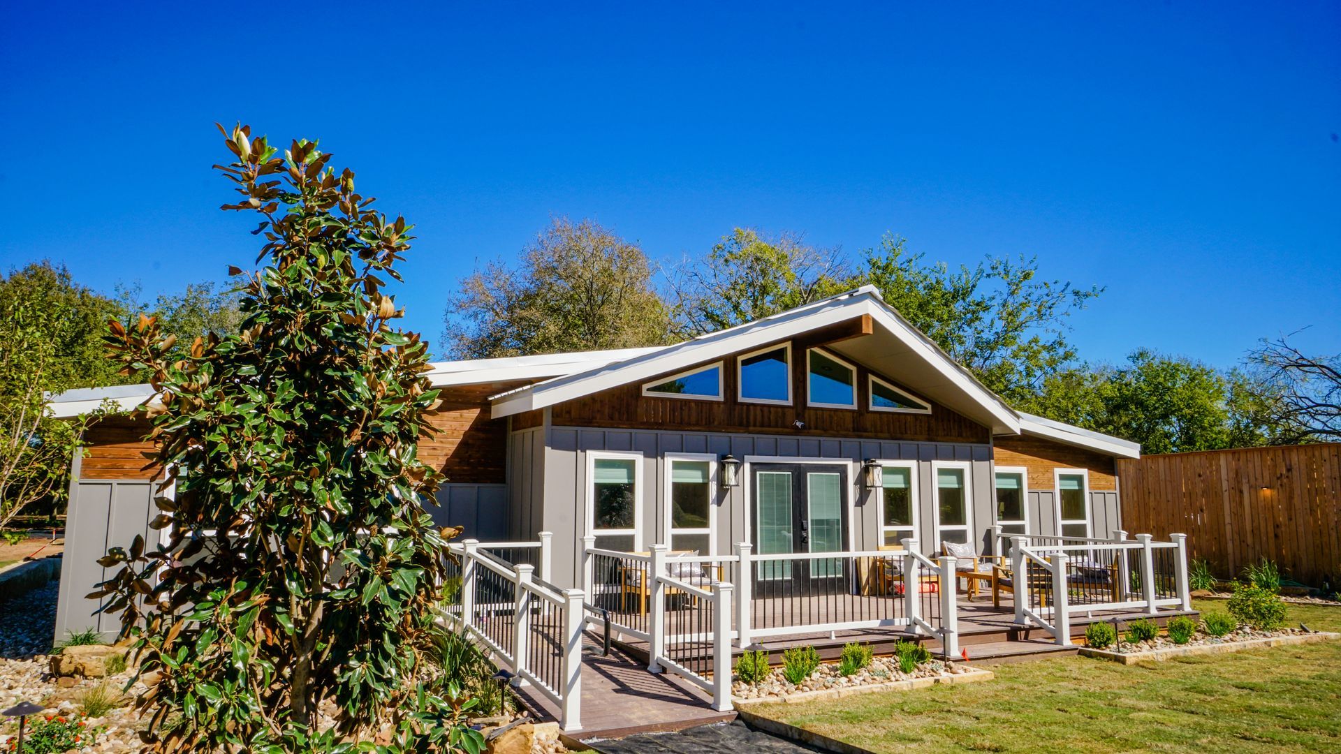 Cottage with gray and brown exterior, wooden deck, glass door, and windows under a blue sky.