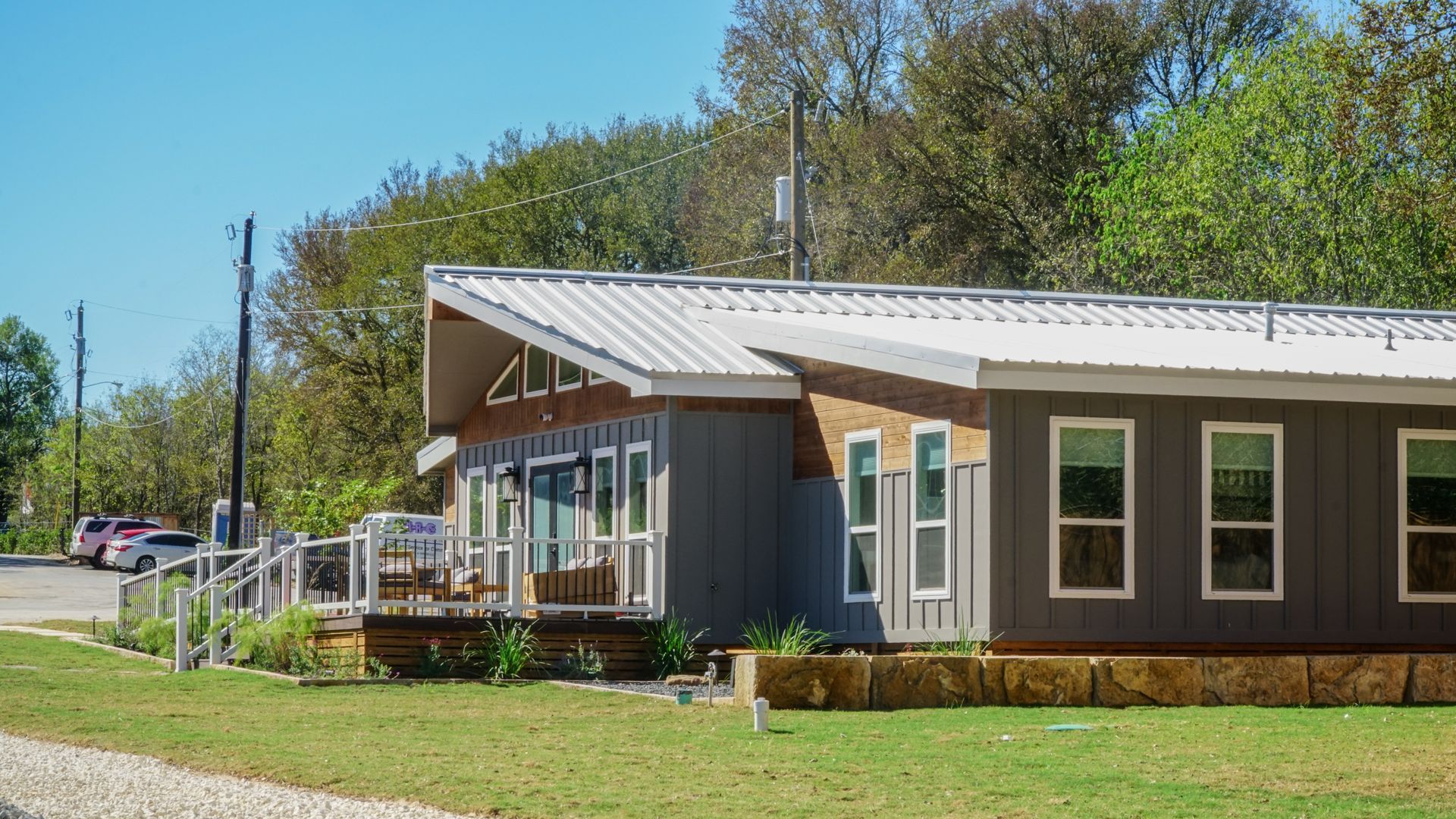 Gray building with a sloped metal roof, windows, and a wooden deck, set against a green lawn and trees.
