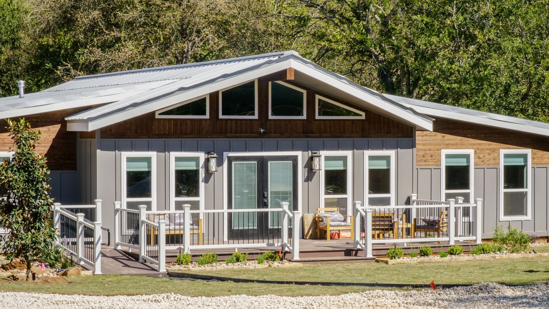 Gray and brown house with a white railing and multiple windows, in a grassy setting.