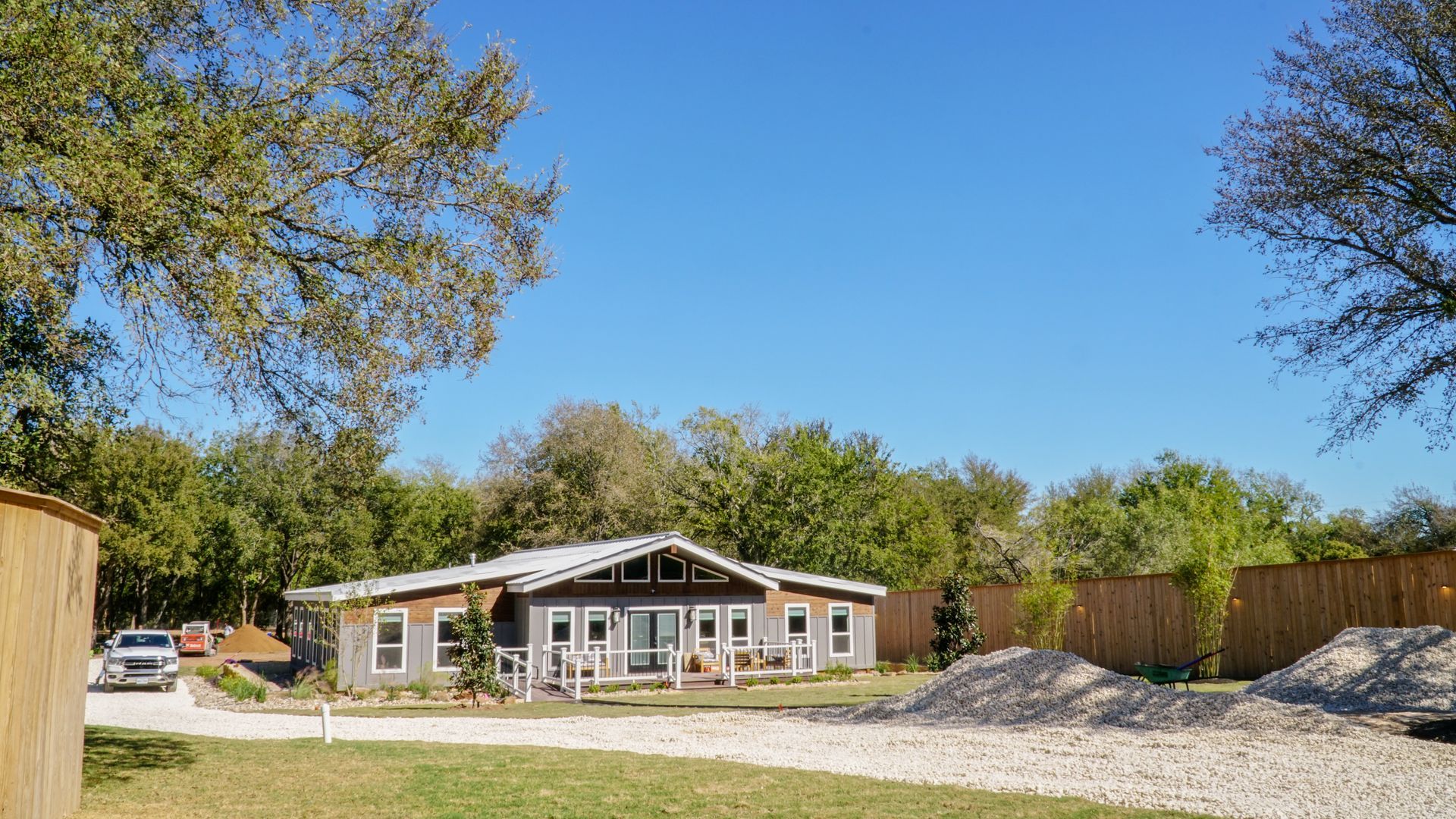 Gray house with white trim under a clear blue sky, surrounded by trees and gravel.
