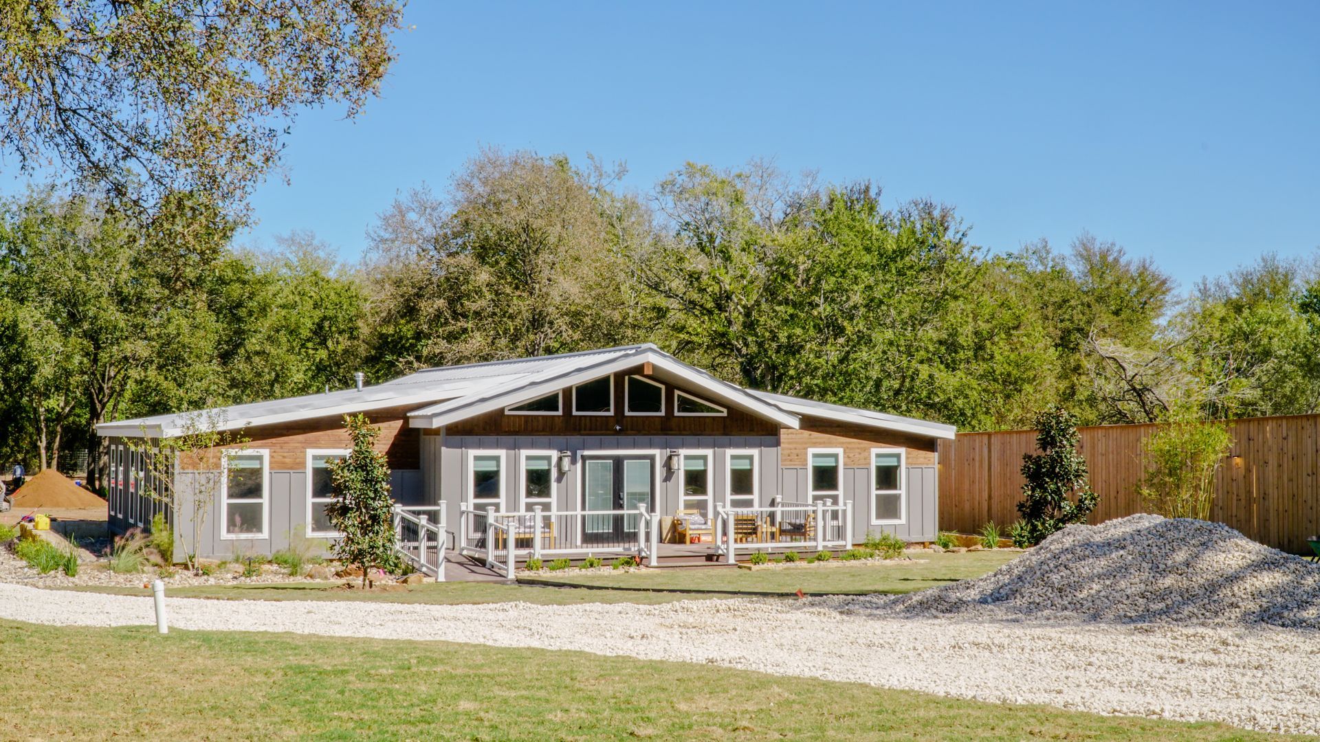 Gray and brown ranch-style house with a gravel driveway, surrounded by trees under a blue sky.