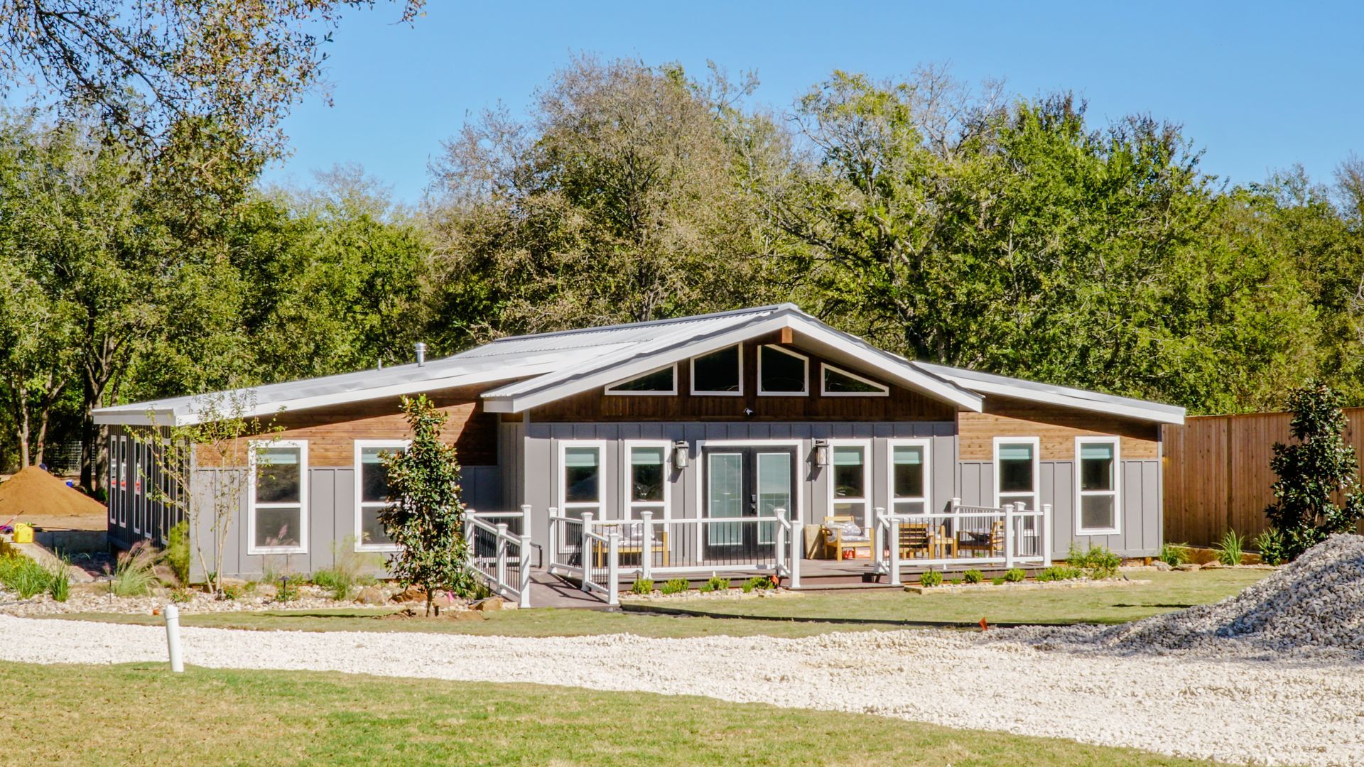 A modern gray and brown house with a large open front porch and a gravel driveway in a wooded area.