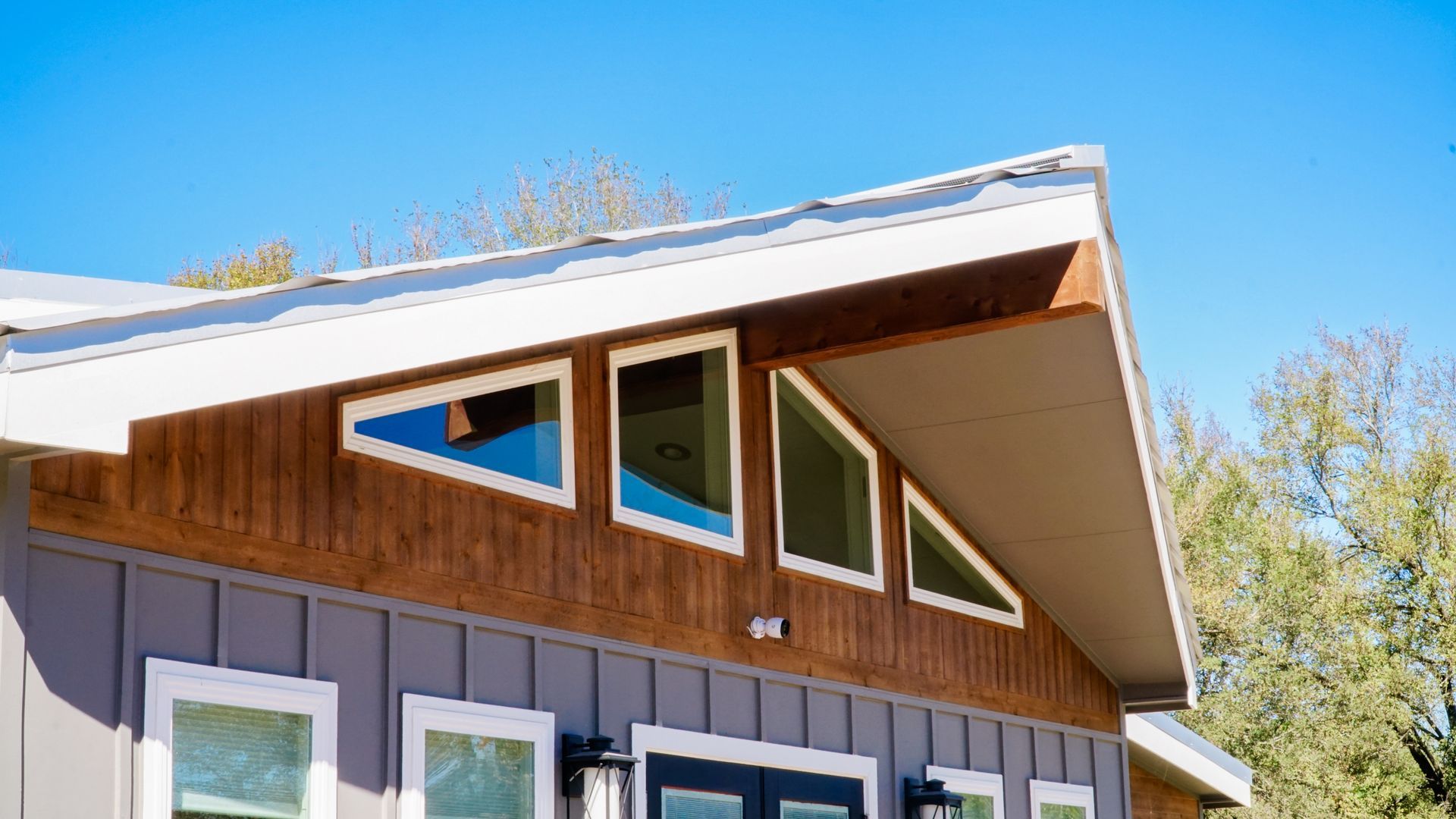 Exterior of a house with gray siding, wooden accents, and triangular windows under a blue sky.