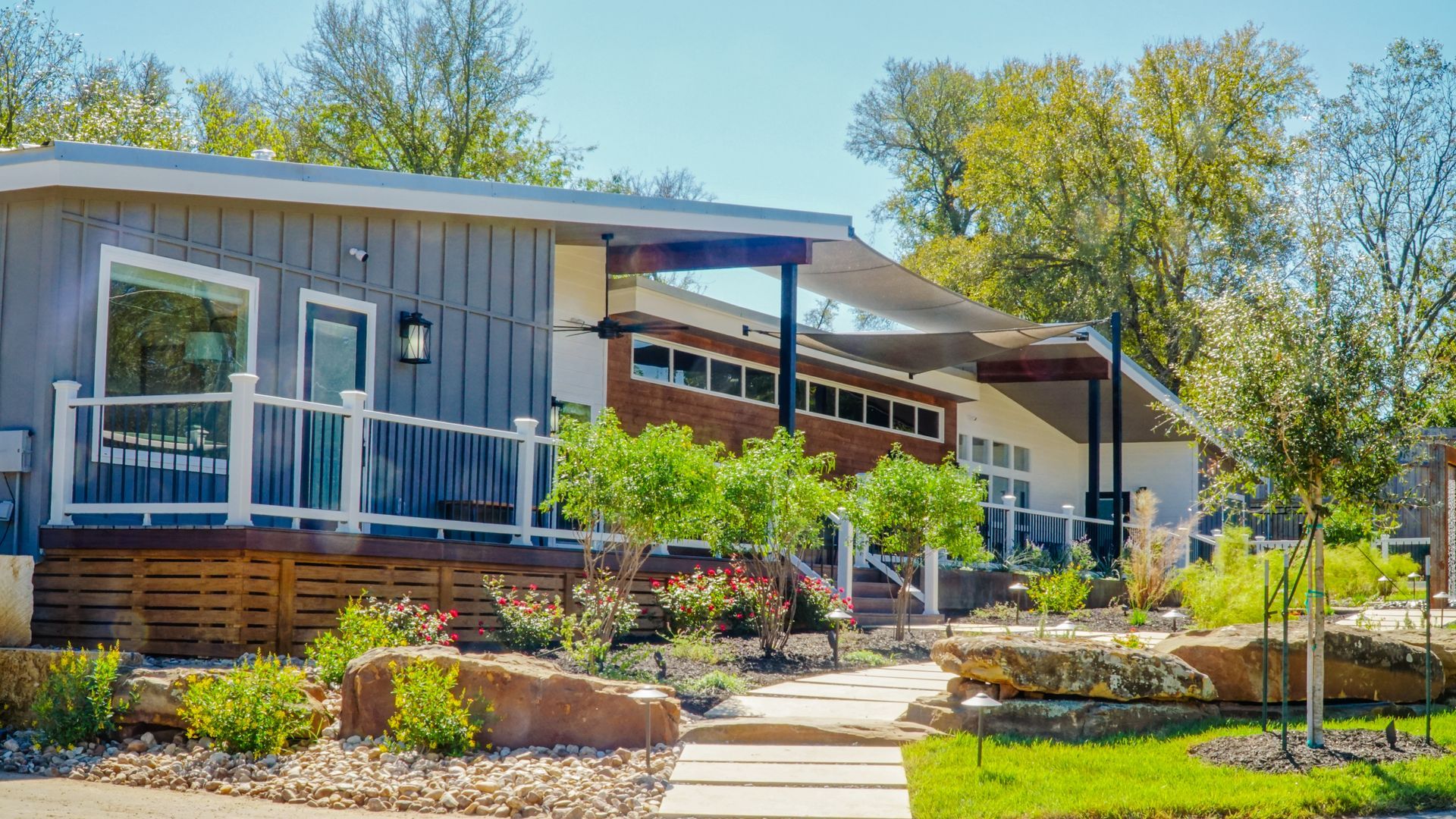 Modern, low-slung building with gray siding, wooden deck, landscaping, and shade sail.