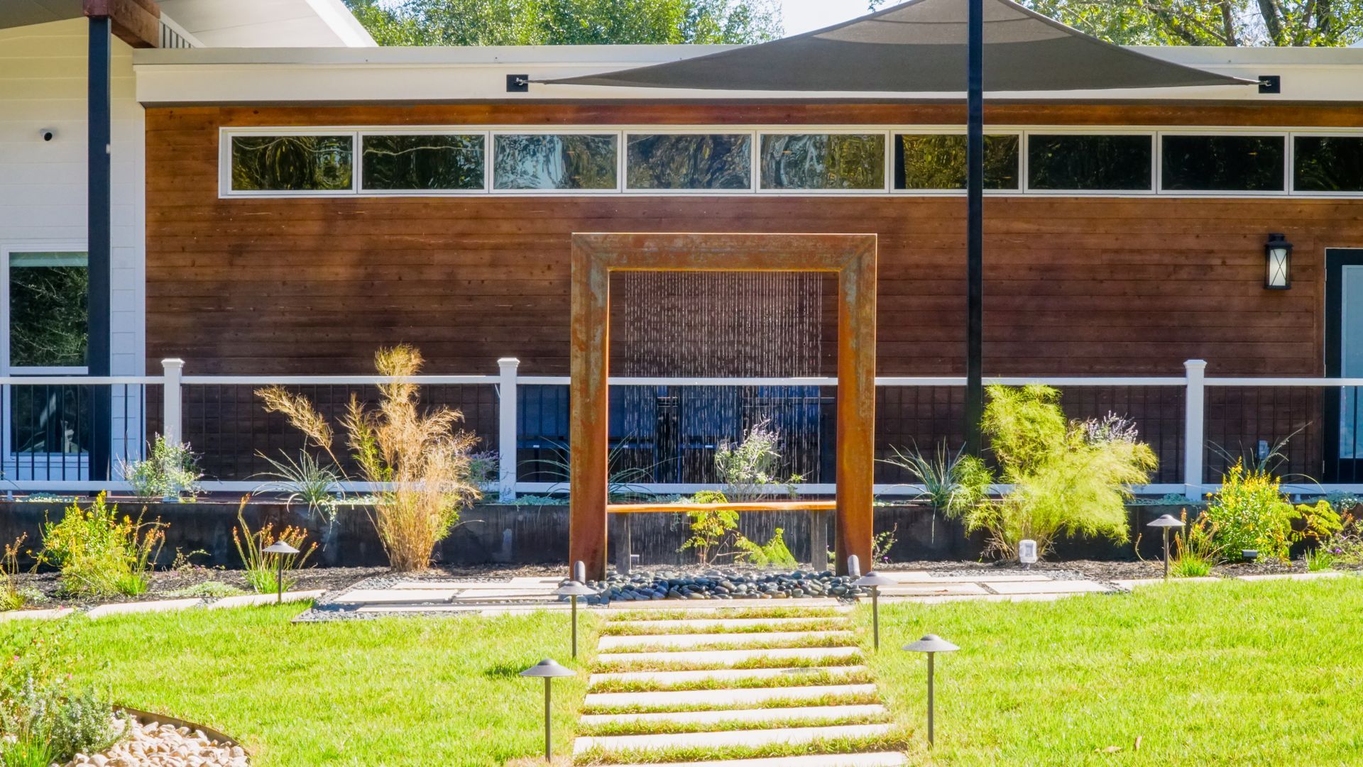 A modern wooden water feature with flowing water in front of a house with a wood exterior and windows.