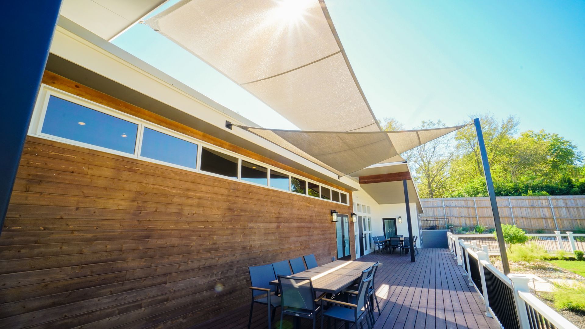 A modern deck with a large shade sail and a long table with chairs. Sunny outdoor setting.