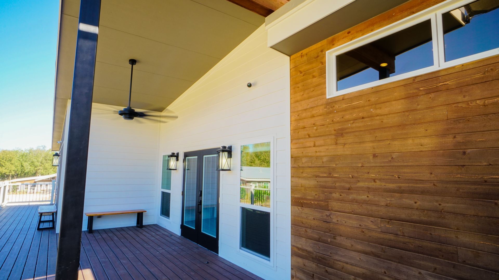 Exterior view of a modern house with a deck. White siding, wood panels, black doors, and a ceiling fan.