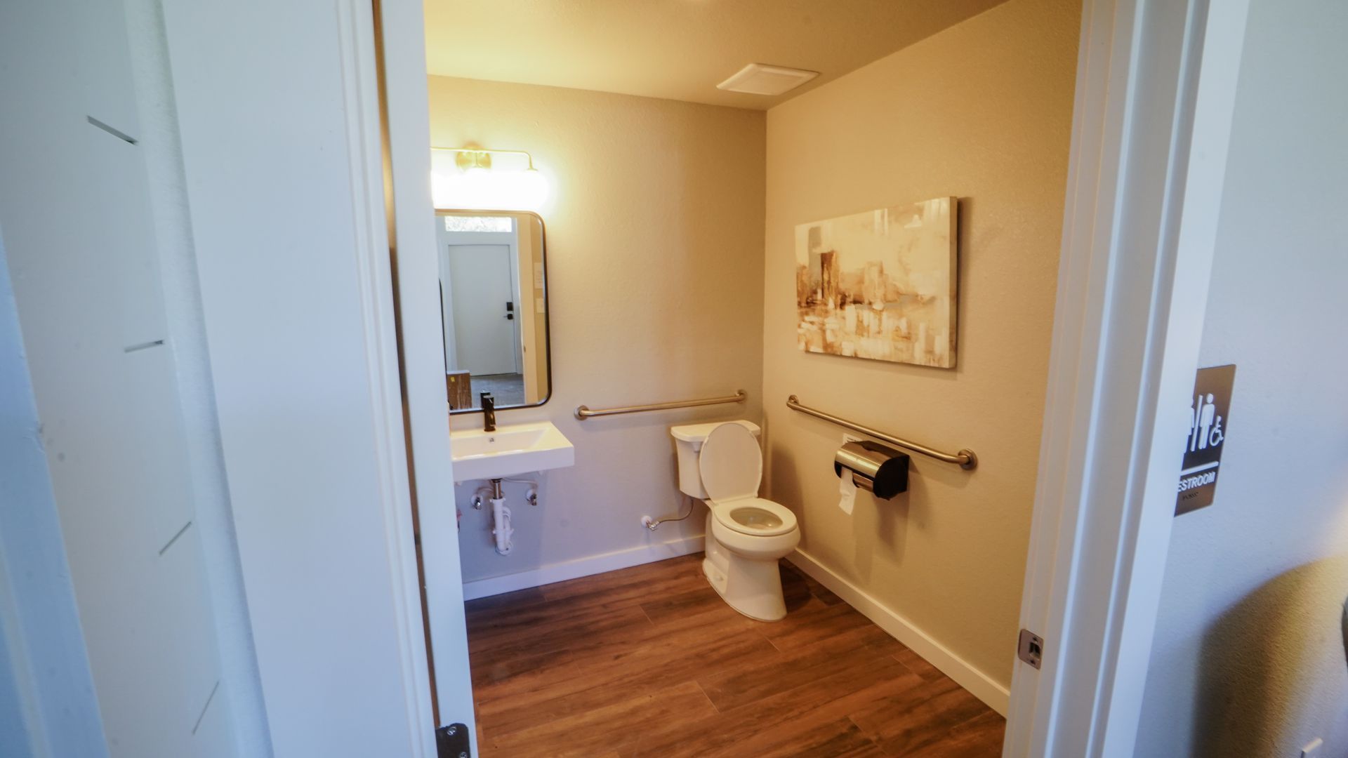 A small bathroom with a toilet, sink, mirror, and grab bars. Features wood-look flooring and light-colored walls.