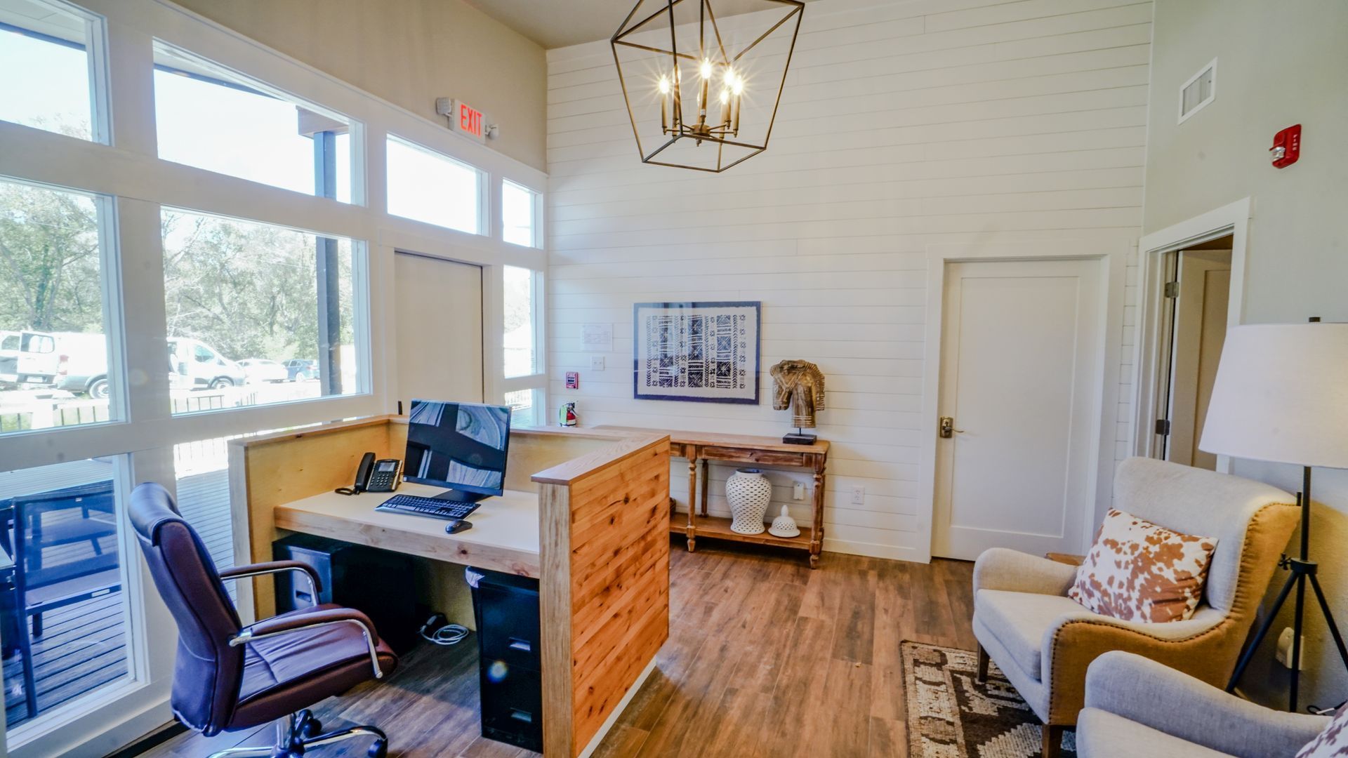 Reception area with desk, two armchairs, large windows, chandelier, and artwork.