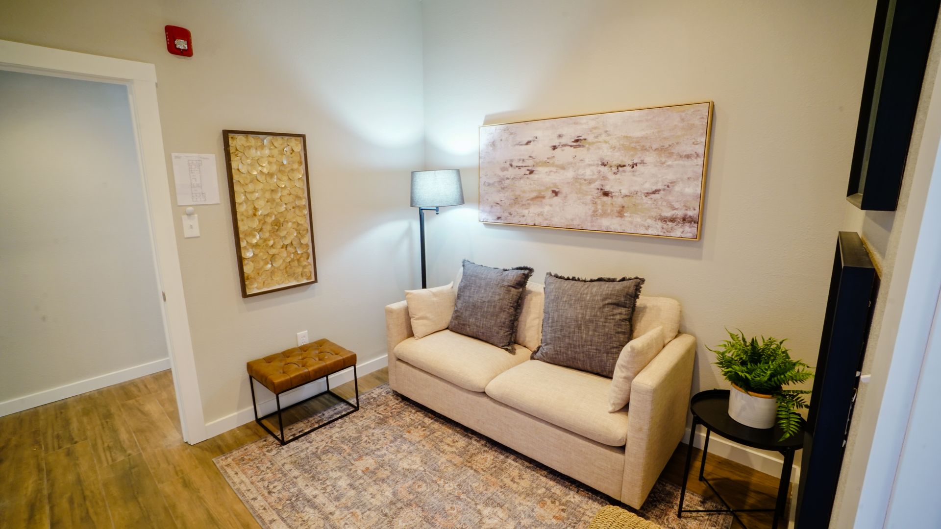 Cozy seating area with a sofa, rug, small tables, artwork, and a floor lamp in a neutral-toned room.