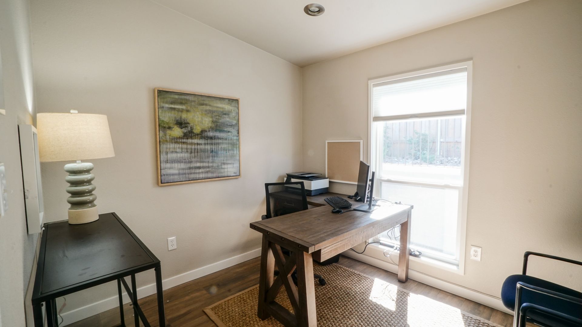 Home office with desk, artwork, window, lamp, and black side table.
