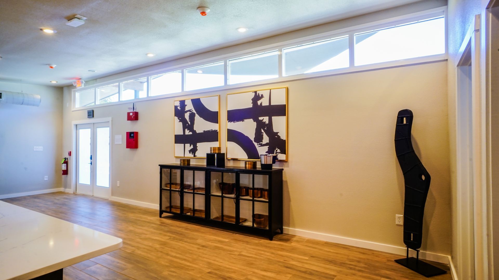Modern room with abstract art, shelving unit, sculpture, and large windows. Beige walls, wooden floor.