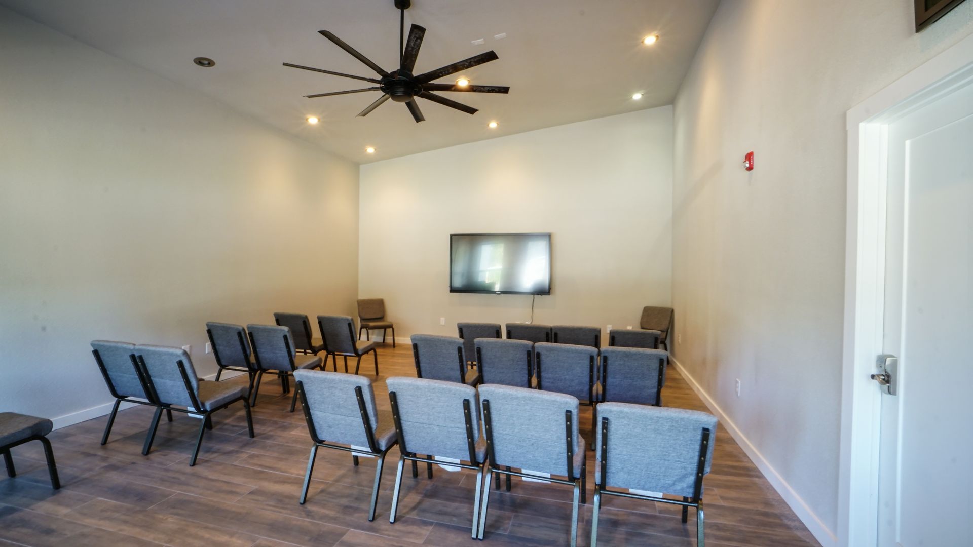 Empty, modern room with rows of gray chairs facing a mounted TV screen. Ceiling fan hangs above.