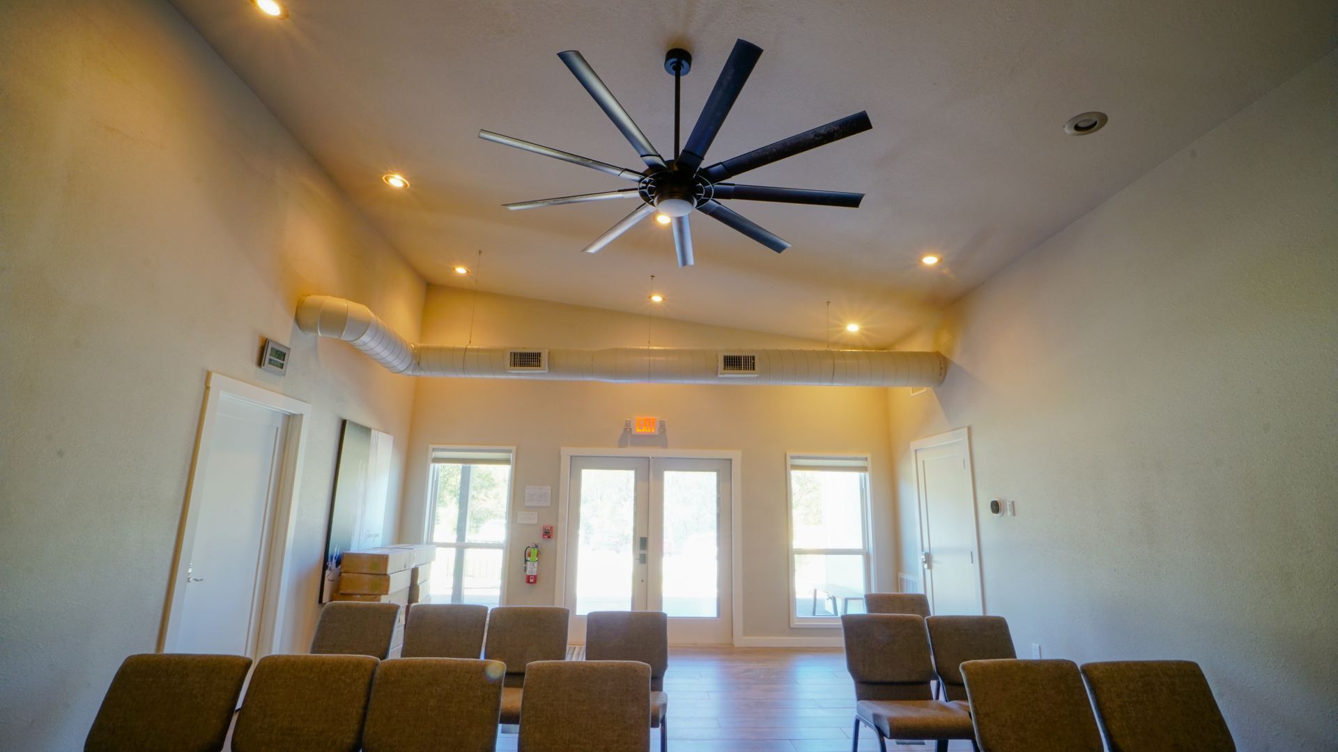 Empty meeting room with ceiling fan, recessed lighting, and rows of chairs facing windows.