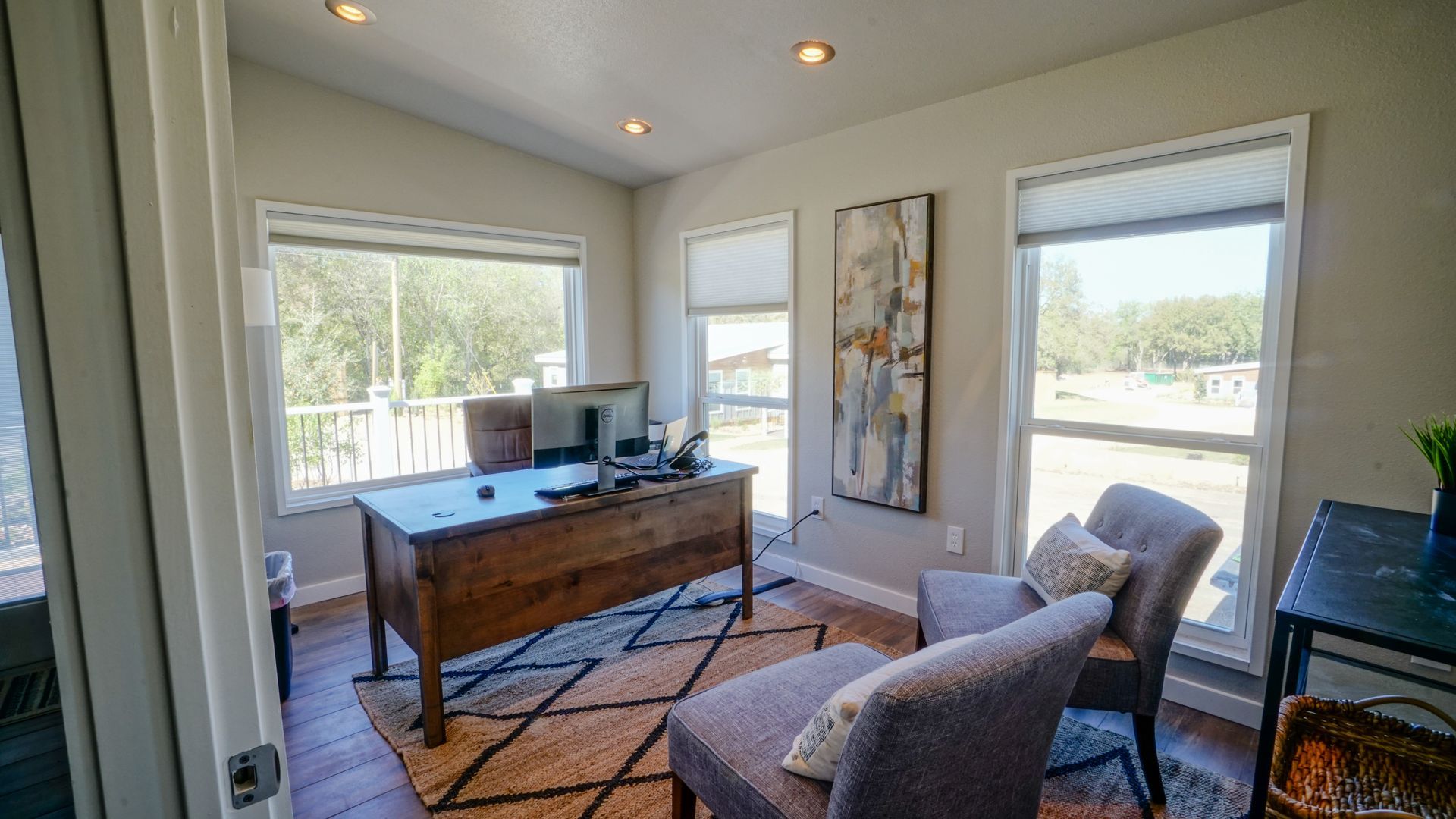 Home office with desk, rug, chairs, artwork, and three windows overlooking a yard.