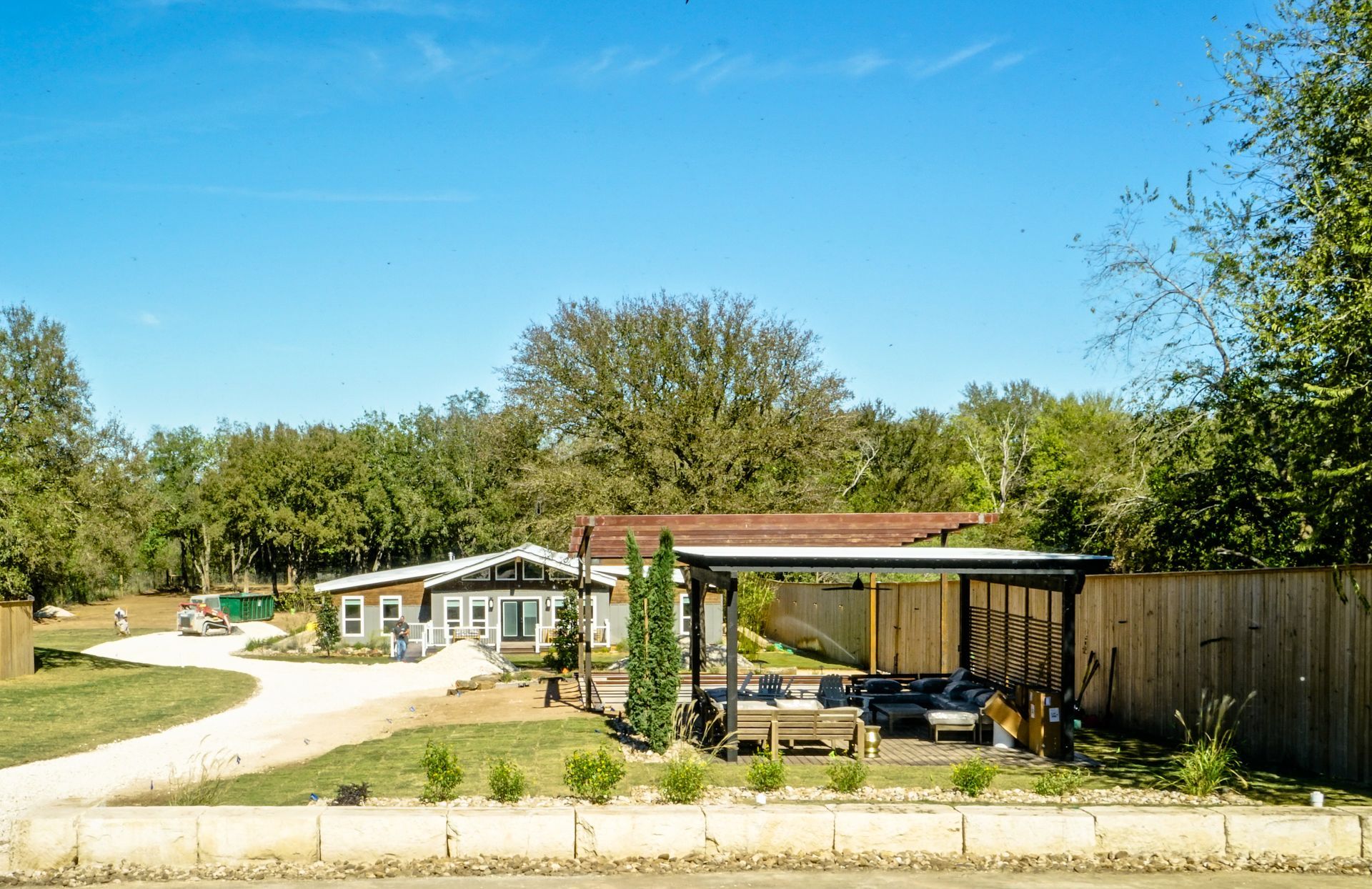 A ranch-style house with a covered carport, gravel driveway, and surrounding trees under a blue sky.
