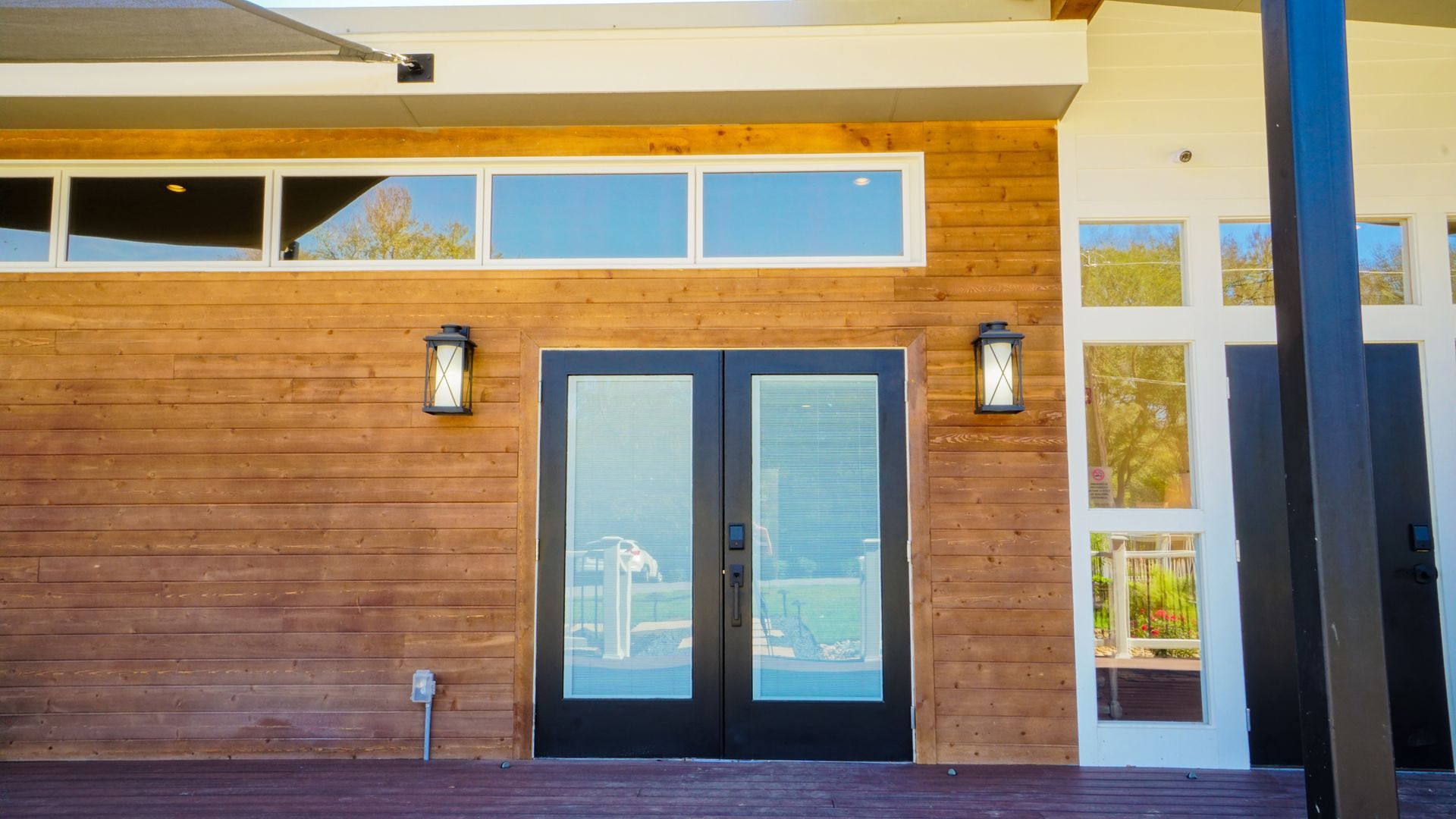 Wooden-paneled building with black-framed glass doors, windows above, and sconce lights.