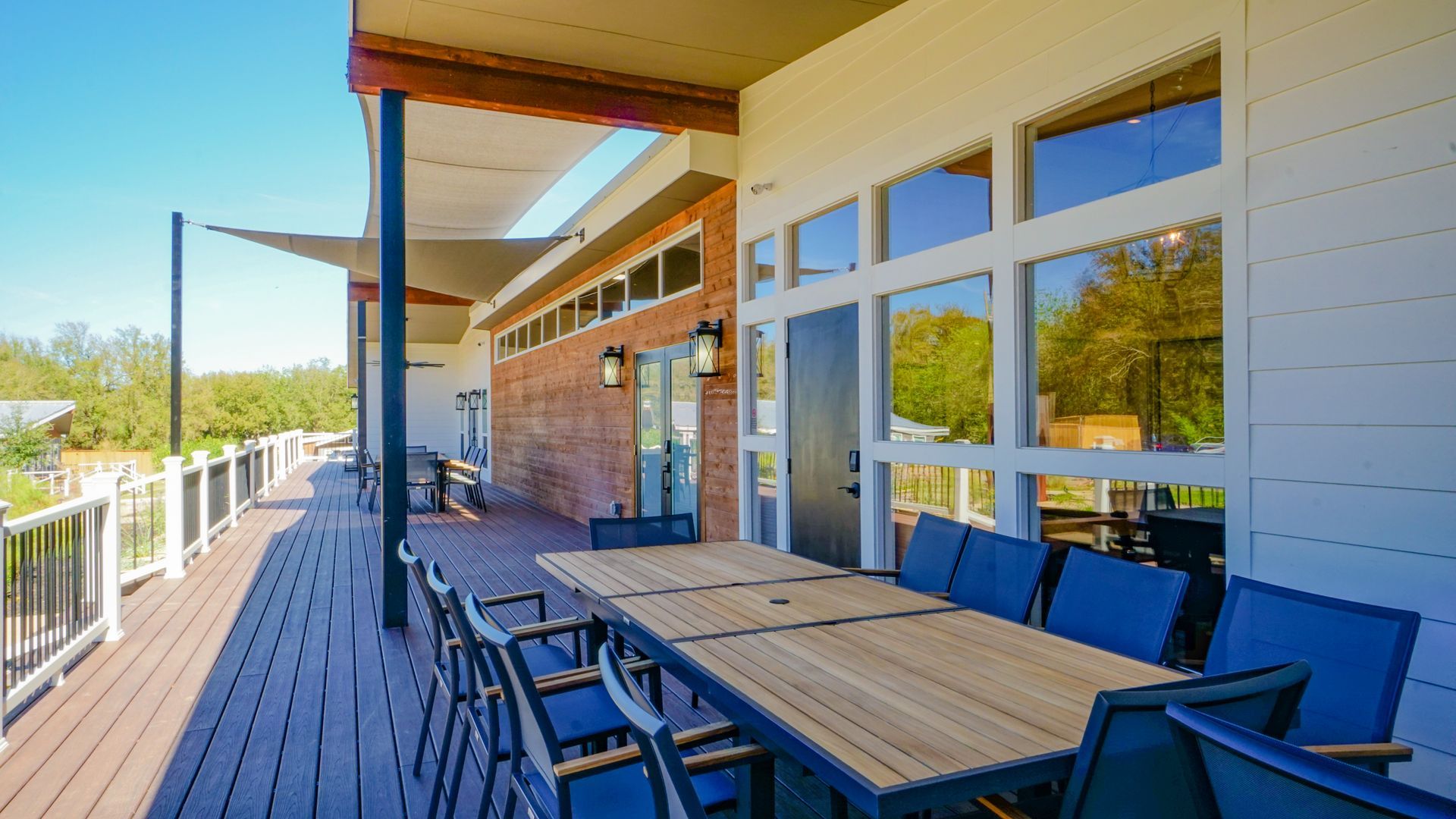Wooden deck with dining table and chairs, adjacent to a building with large windows and brick accent wall, on a sunny day.