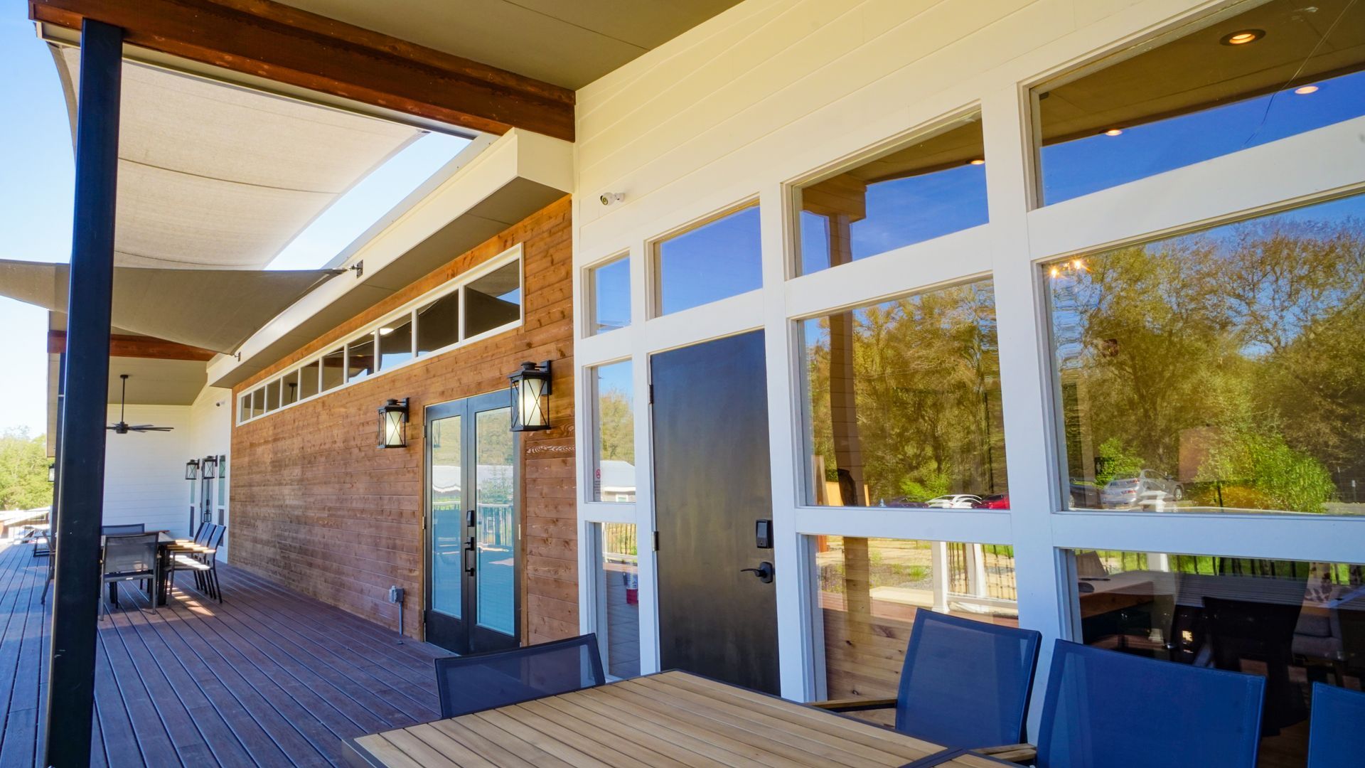 Outdoor deck with tables, chairs, and large windows reflecting trees.  A brick wall and black door are visible.