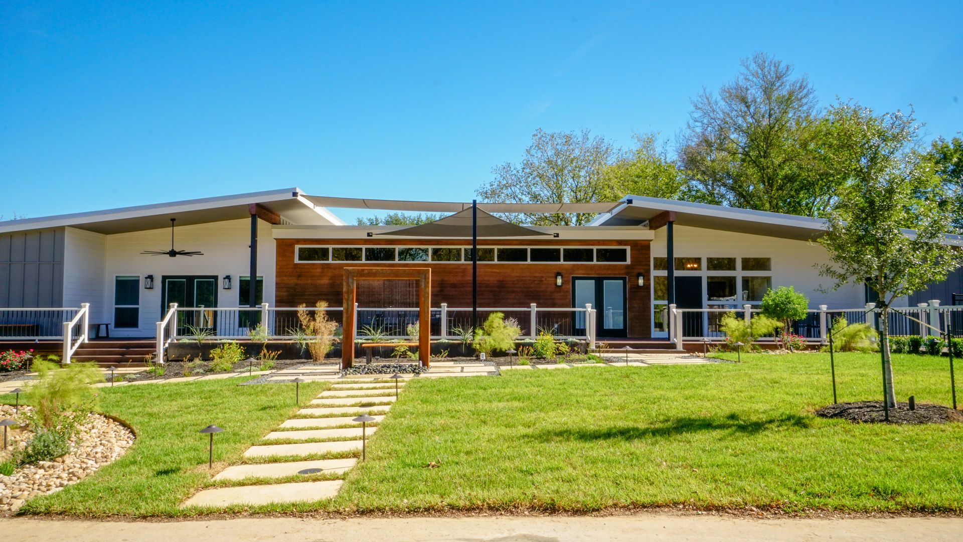 Modern one-story house with a wooden facade, white accents, and a stone path leading to the entrance under a clear blue sky.