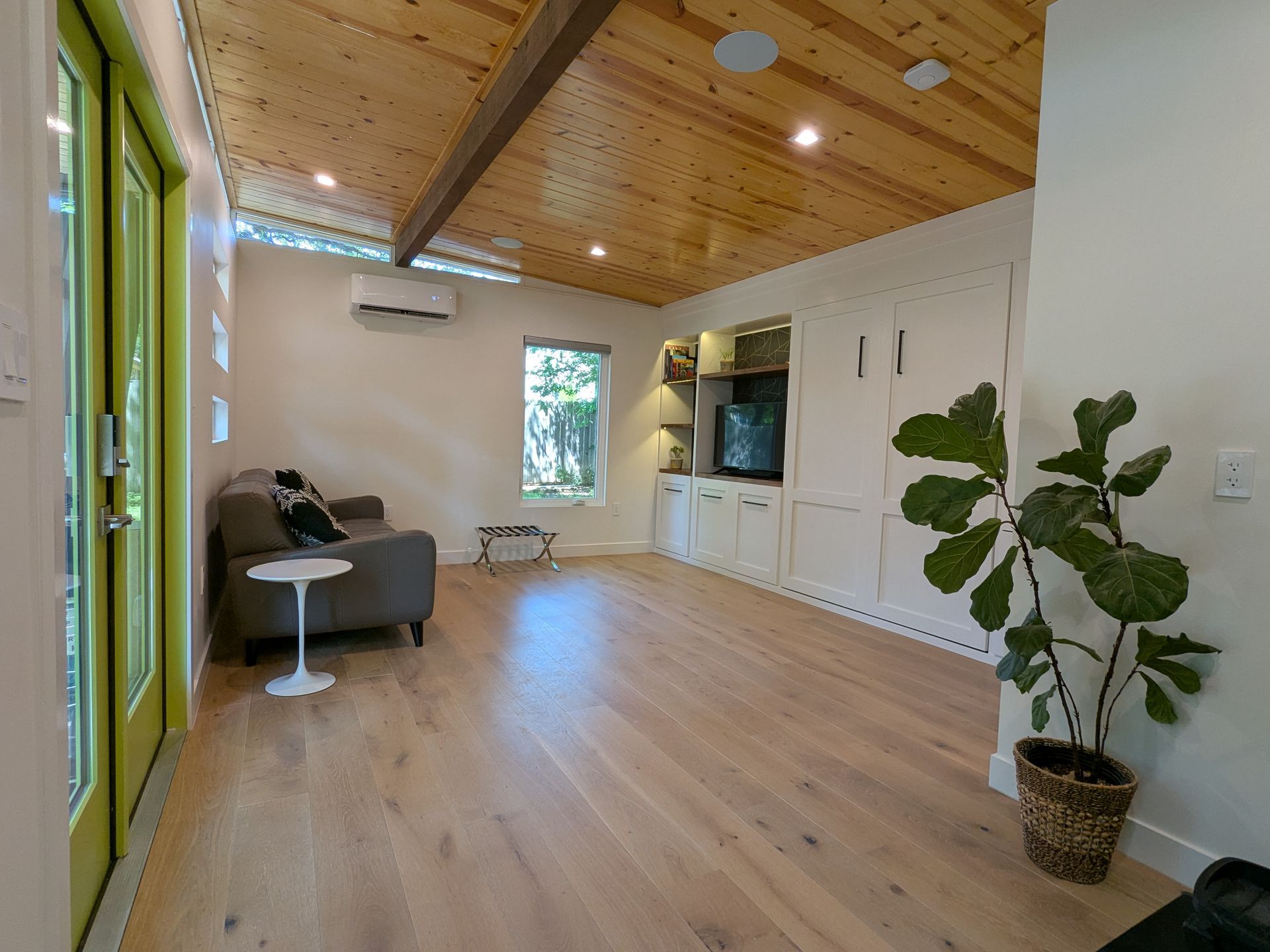 Living room with wood floors, ceiling, and white built-in storage. A green door and a fiddle leaf fig plant are visible.