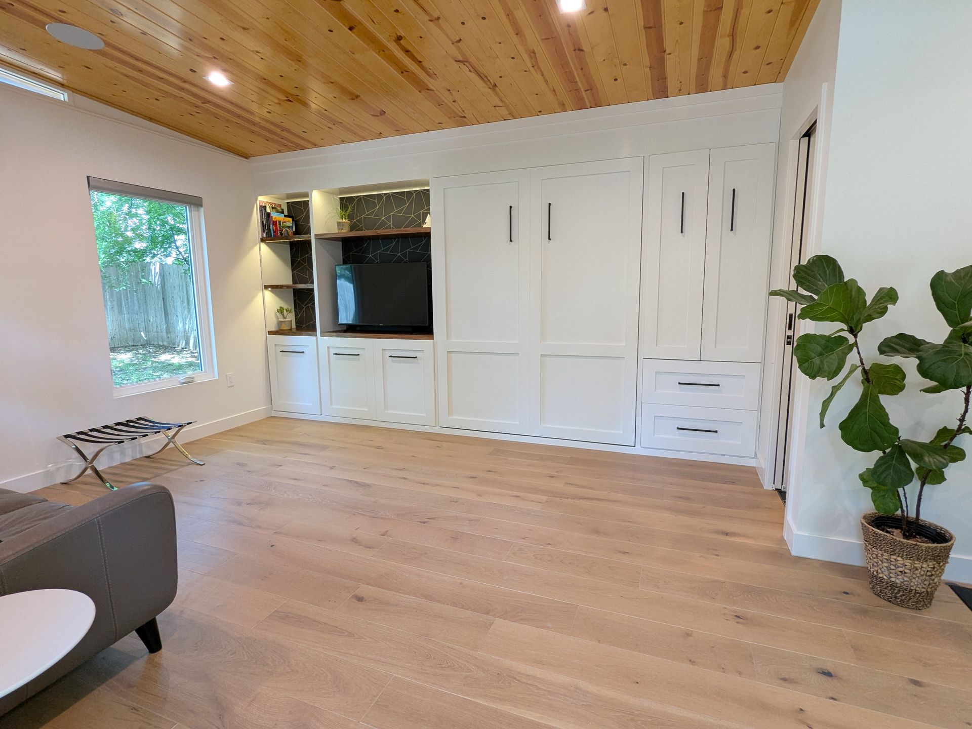 Living room with built-in white cabinets, TV, wooden floor and ceiling, potted plant, and window.