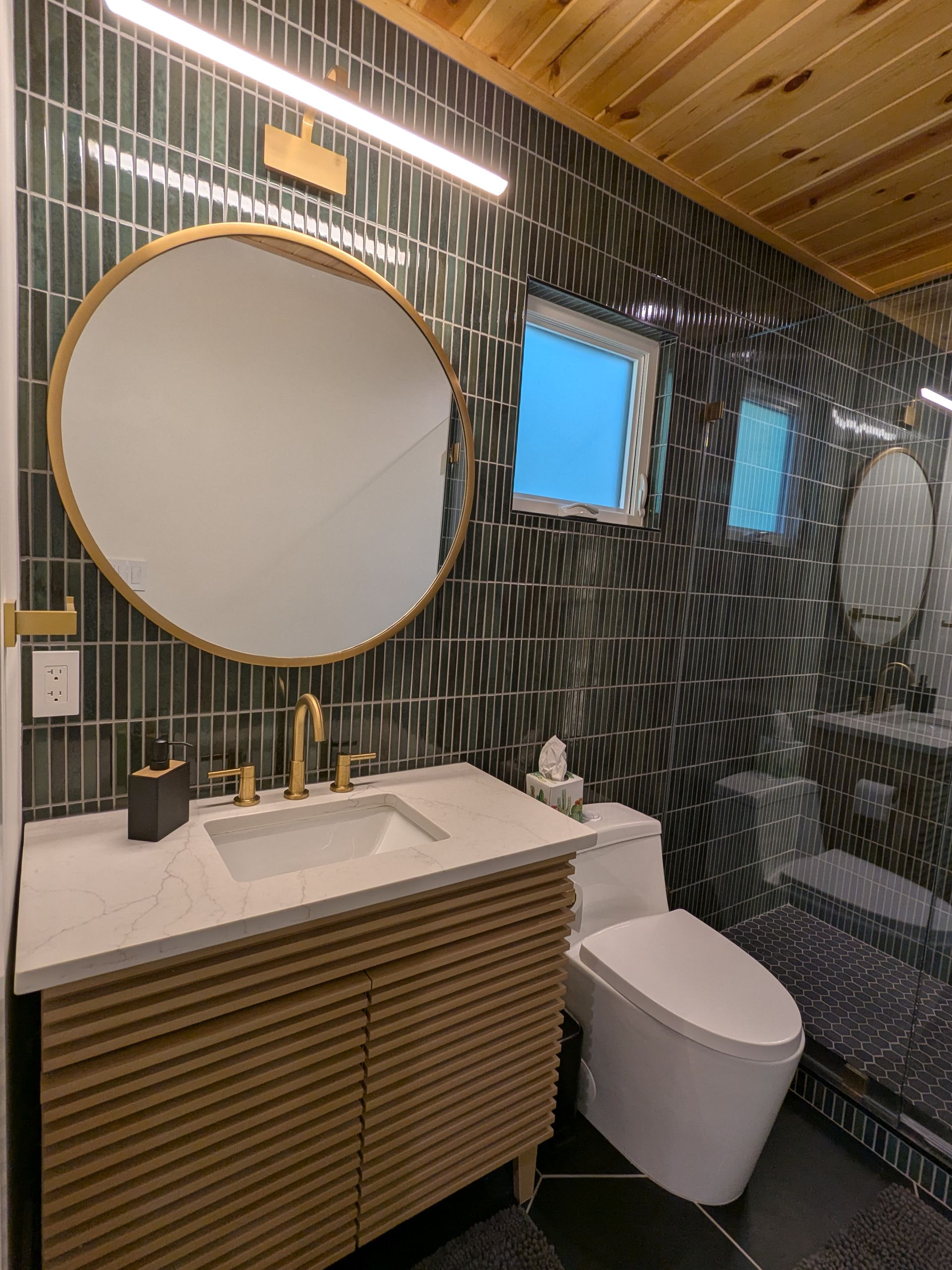 Modern bathroom with a wood vanity, gold fixtures, and patterned black tile.