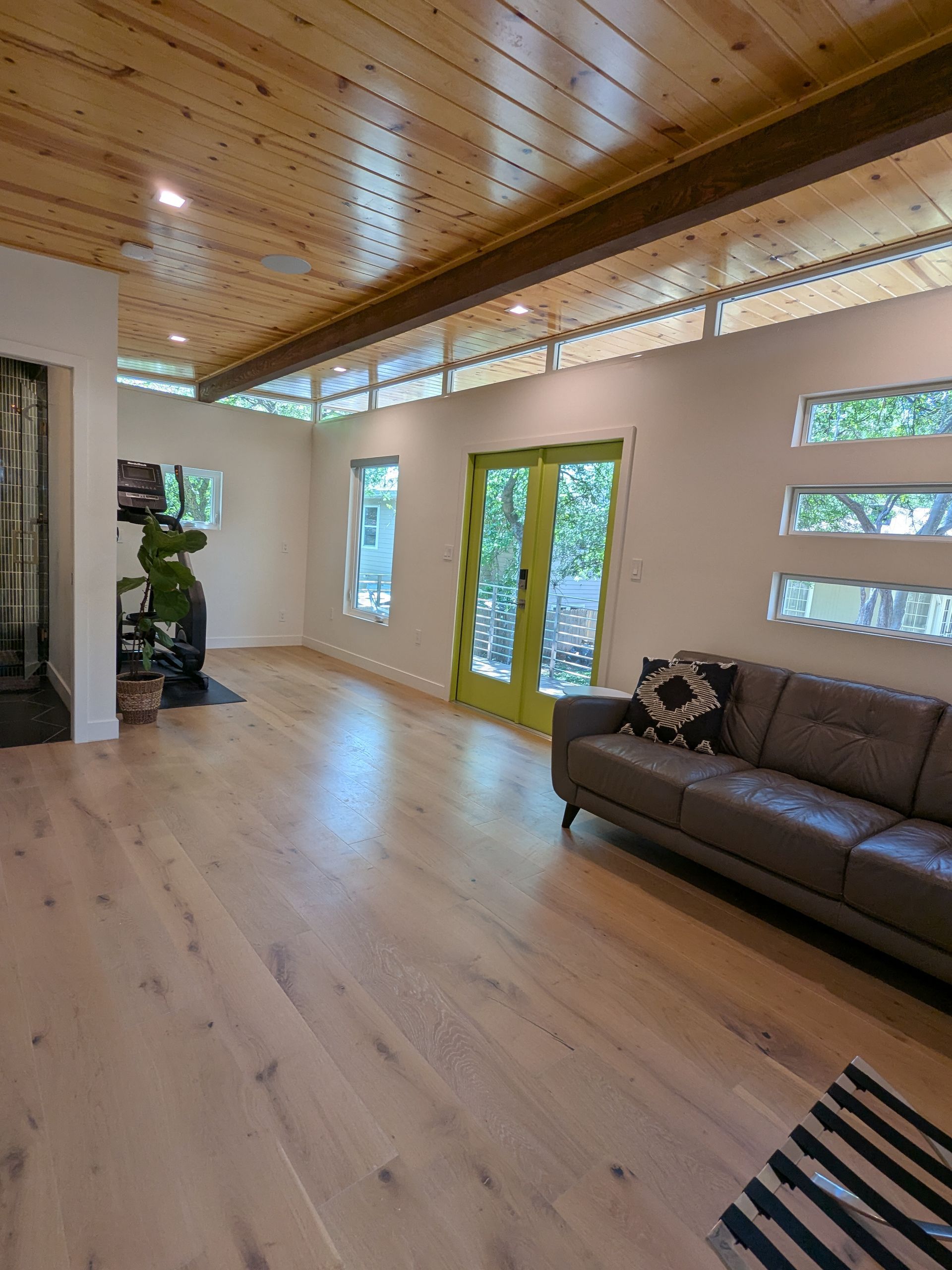 Modern living room with wood floors, light walls, wooden ceiling, and green doors. A grey leather couch is visible.