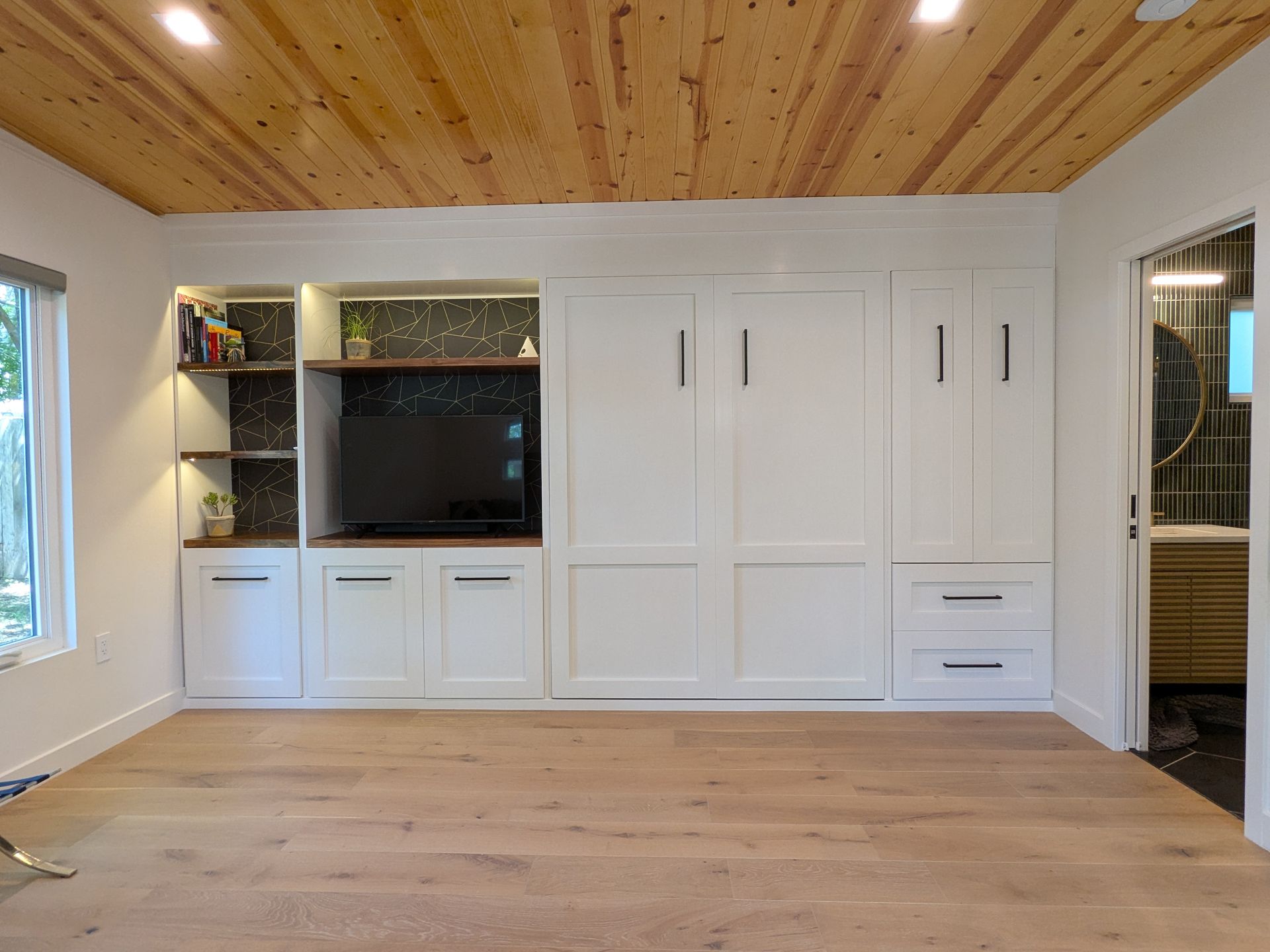A white built-in wall unit with a TV, cabinets, and a fold-out bed in a room with wood floors and ceiling.