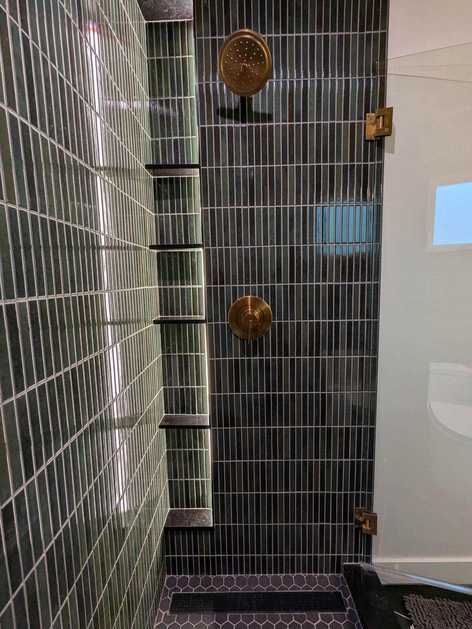 Shower with dark green rectangular tiles, brass fixtures, and built-in shelves.