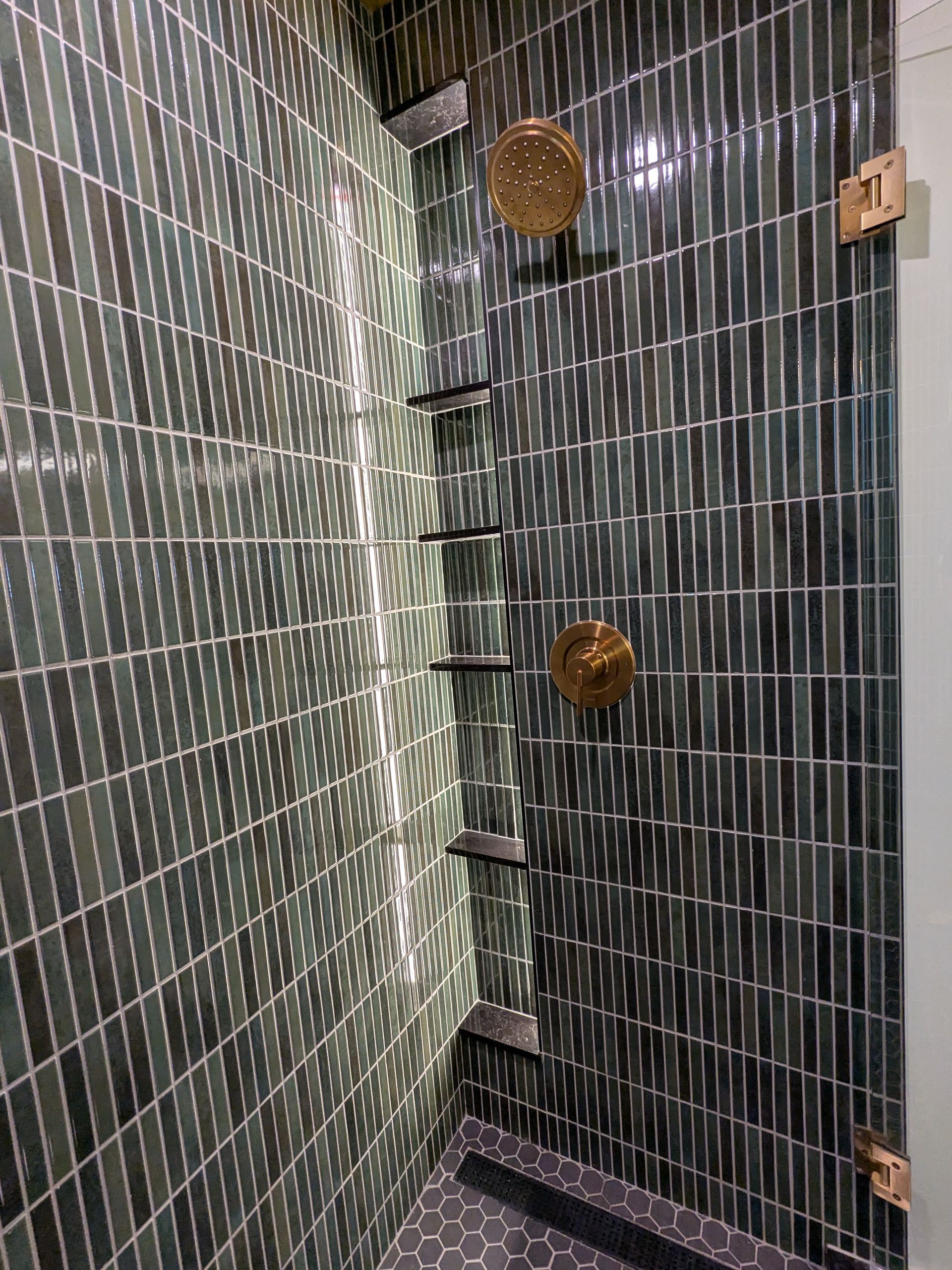 Green tiled shower with built-in shelves, gold fixtures, and dark floor.
