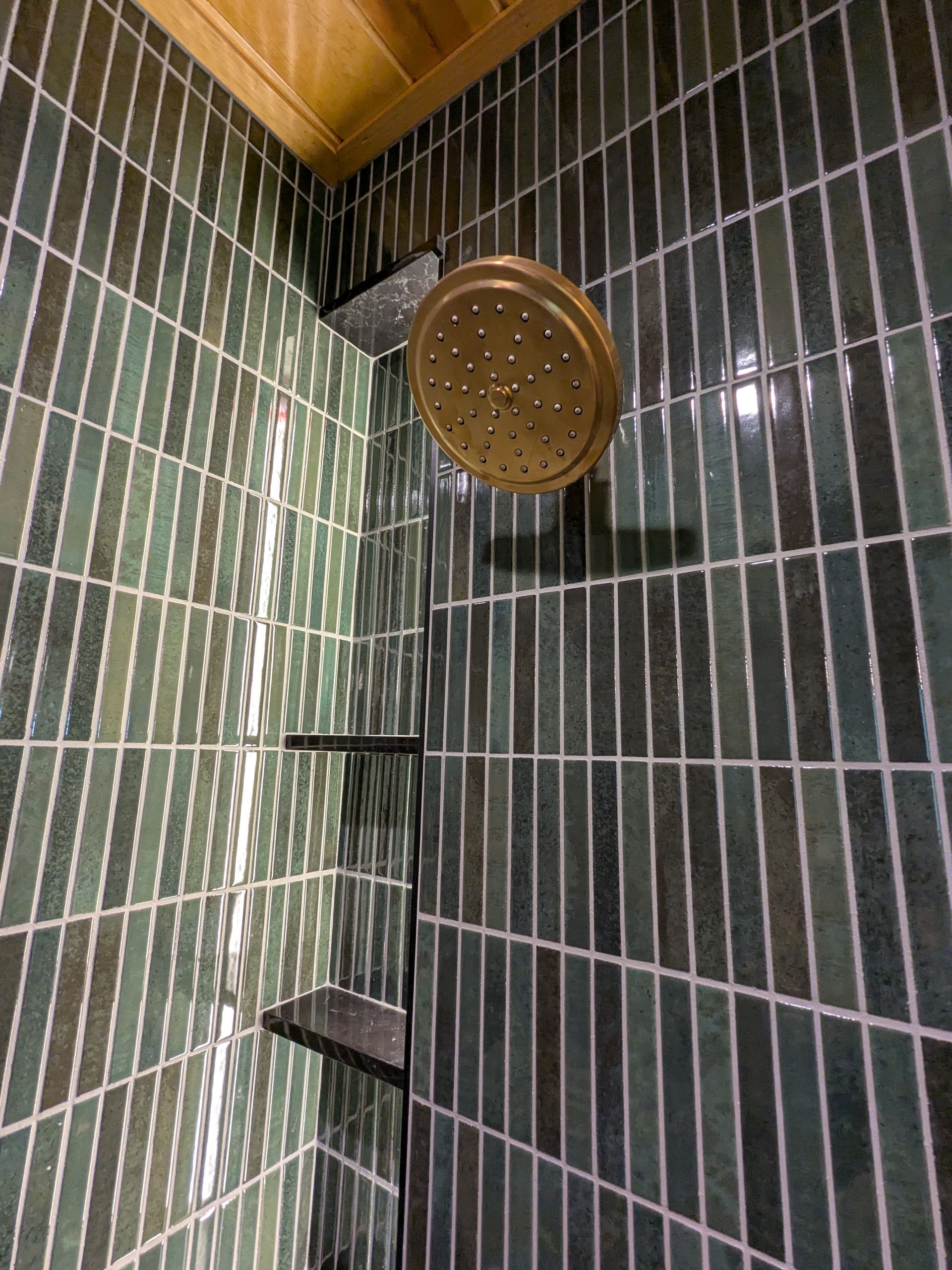 Shower with green rectangular tiles and bronze showerhead. Two dark shelves. Wooden ceiling visible.
