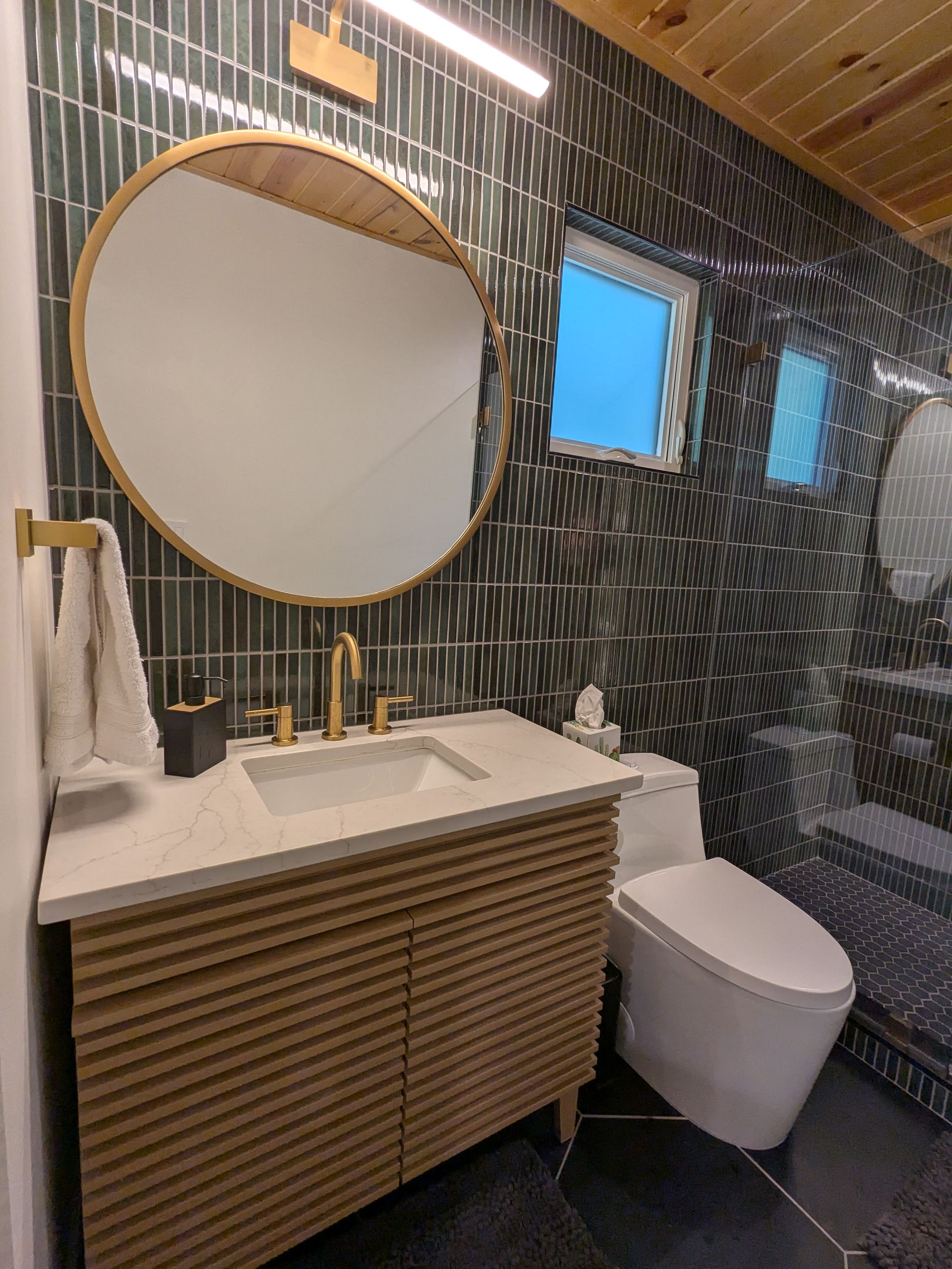 Bathroom with gold fixtures, wood vanity, and patterned tile. Round mirror and small window.