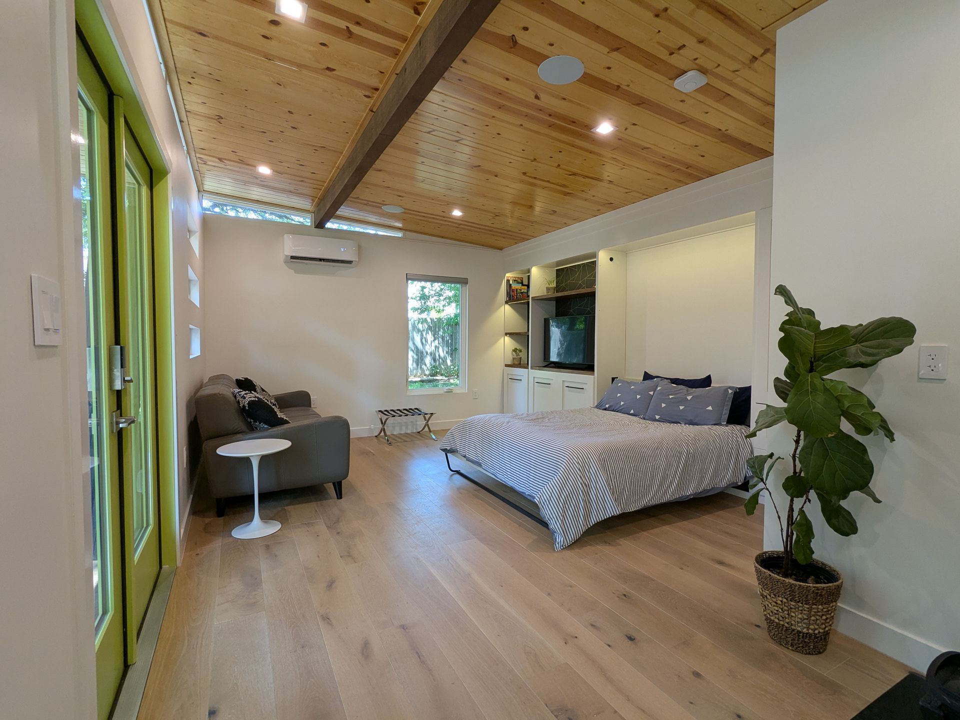 Bright room with wood ceiling, bed, sofa, and a potted plant. Door on left.