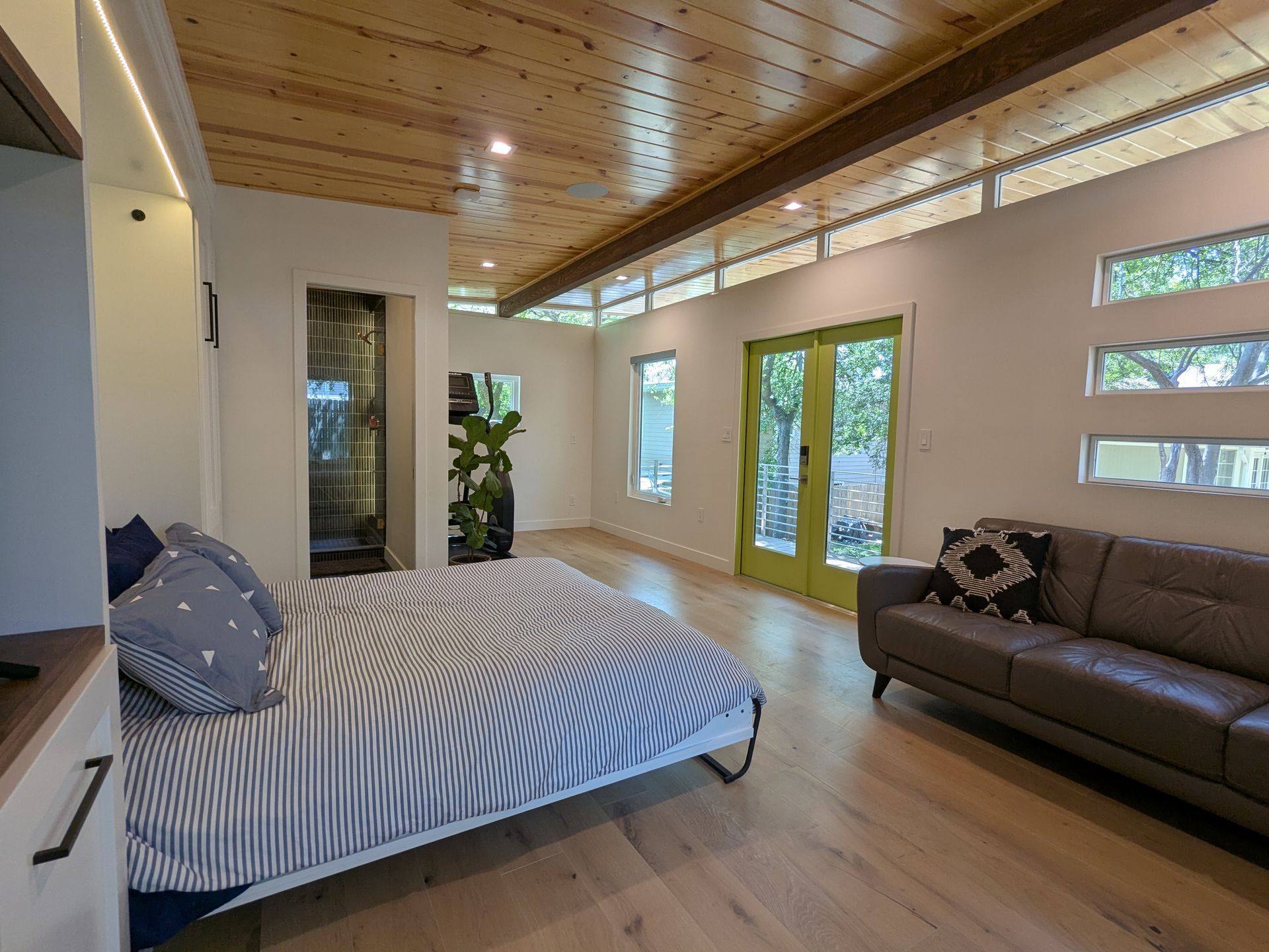 Bedroom with a bed, sofa, wood ceiling, green doors, and large windows.