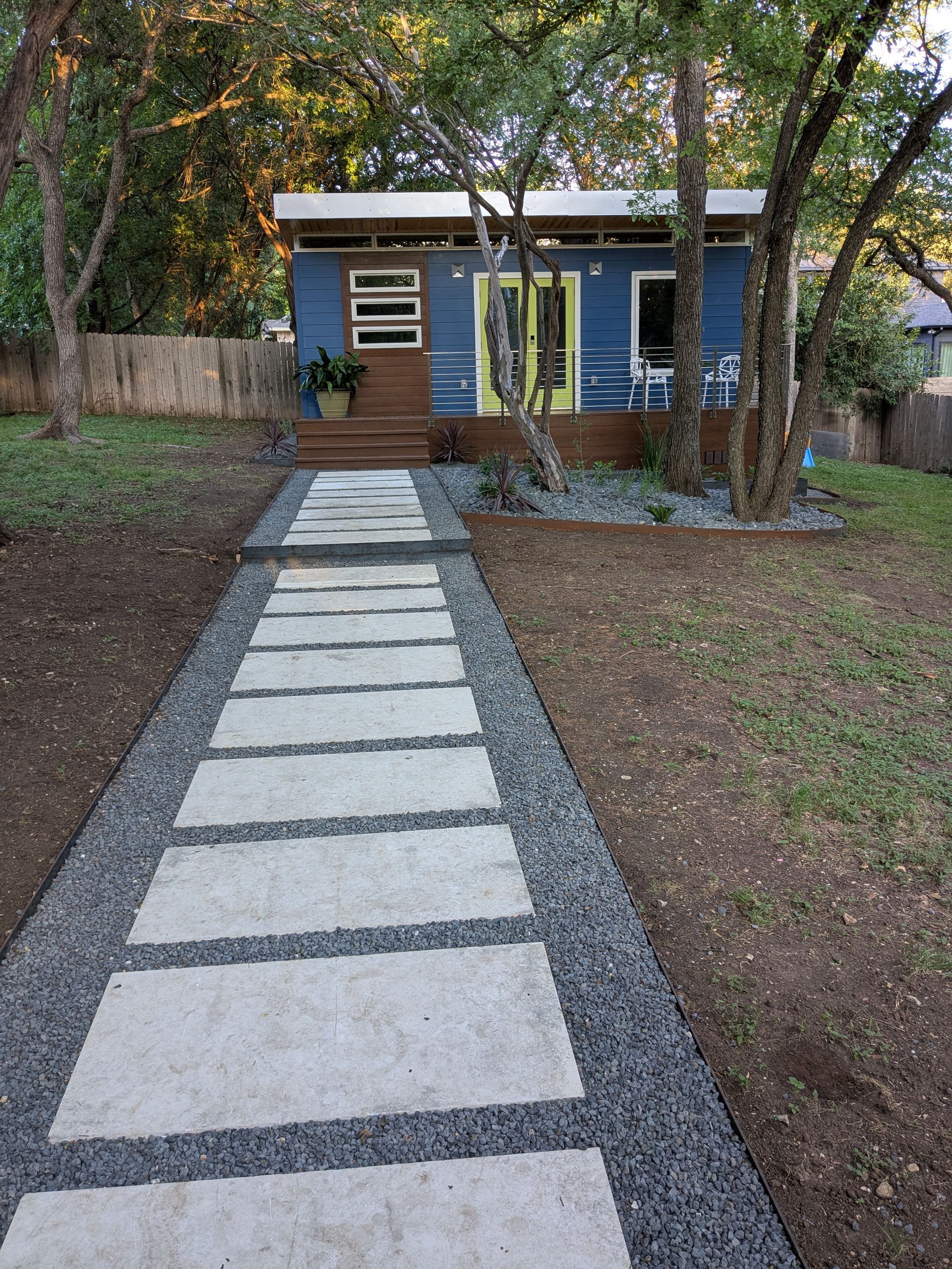 Stone path leads to a small, blue house with a yellow door, surrounded by trees.