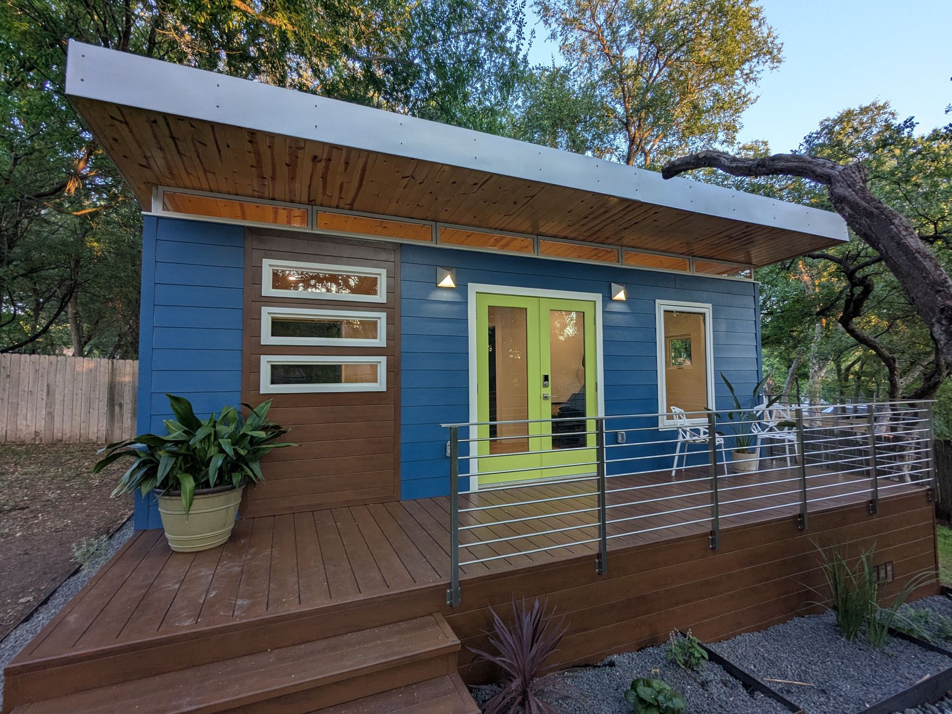 Blue tiny house with wooden deck and green door, under a sloping roof.