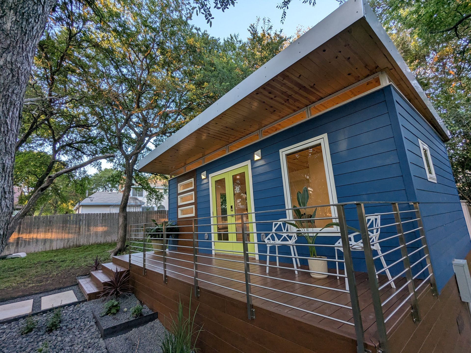 Blue tiny home with wood deck, yellow door, and metal railing in a yard with trees.
