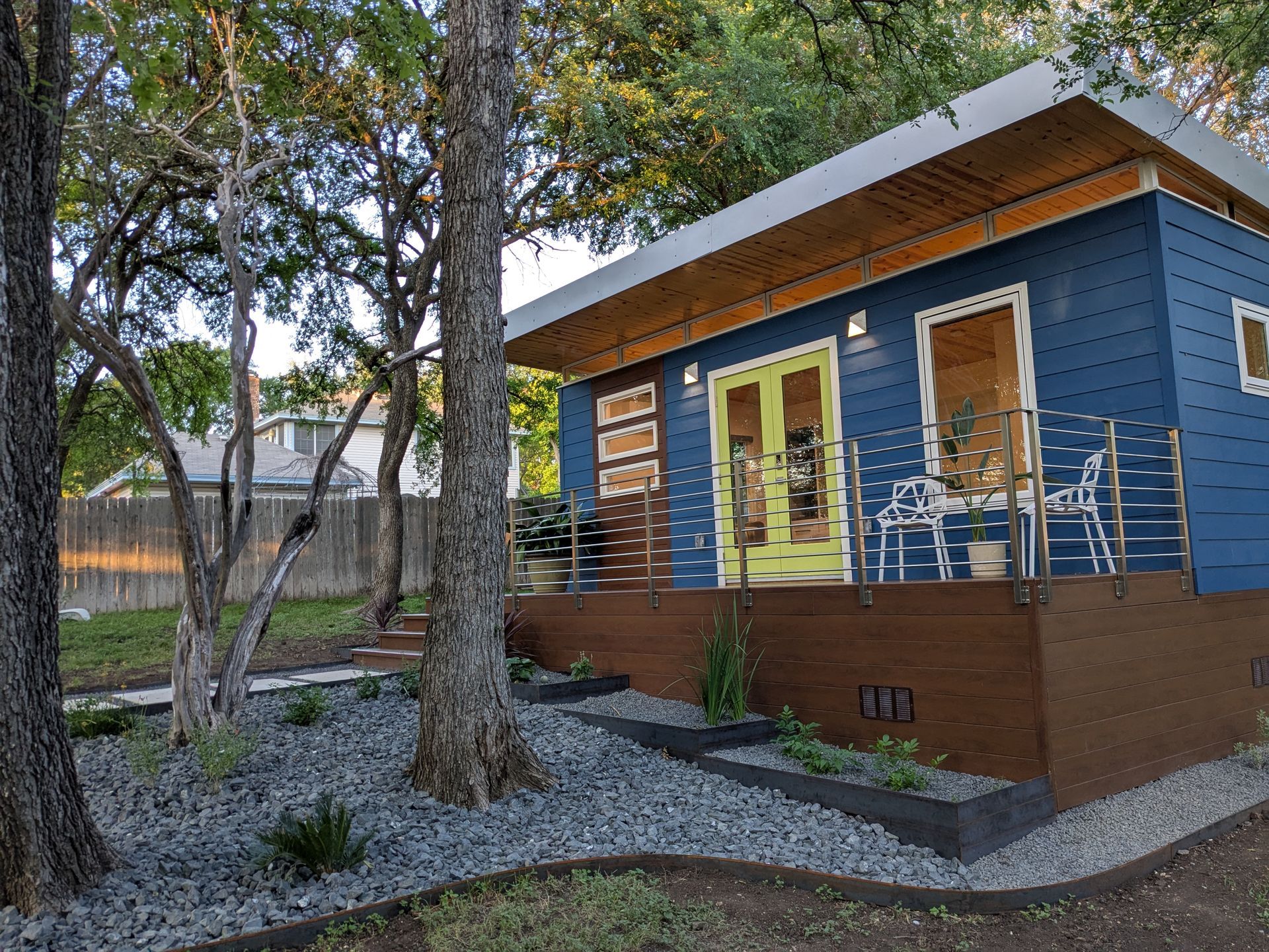 Blue-painted tiny house with yellow doors and wooden deck, surrounded by trees and gravel.