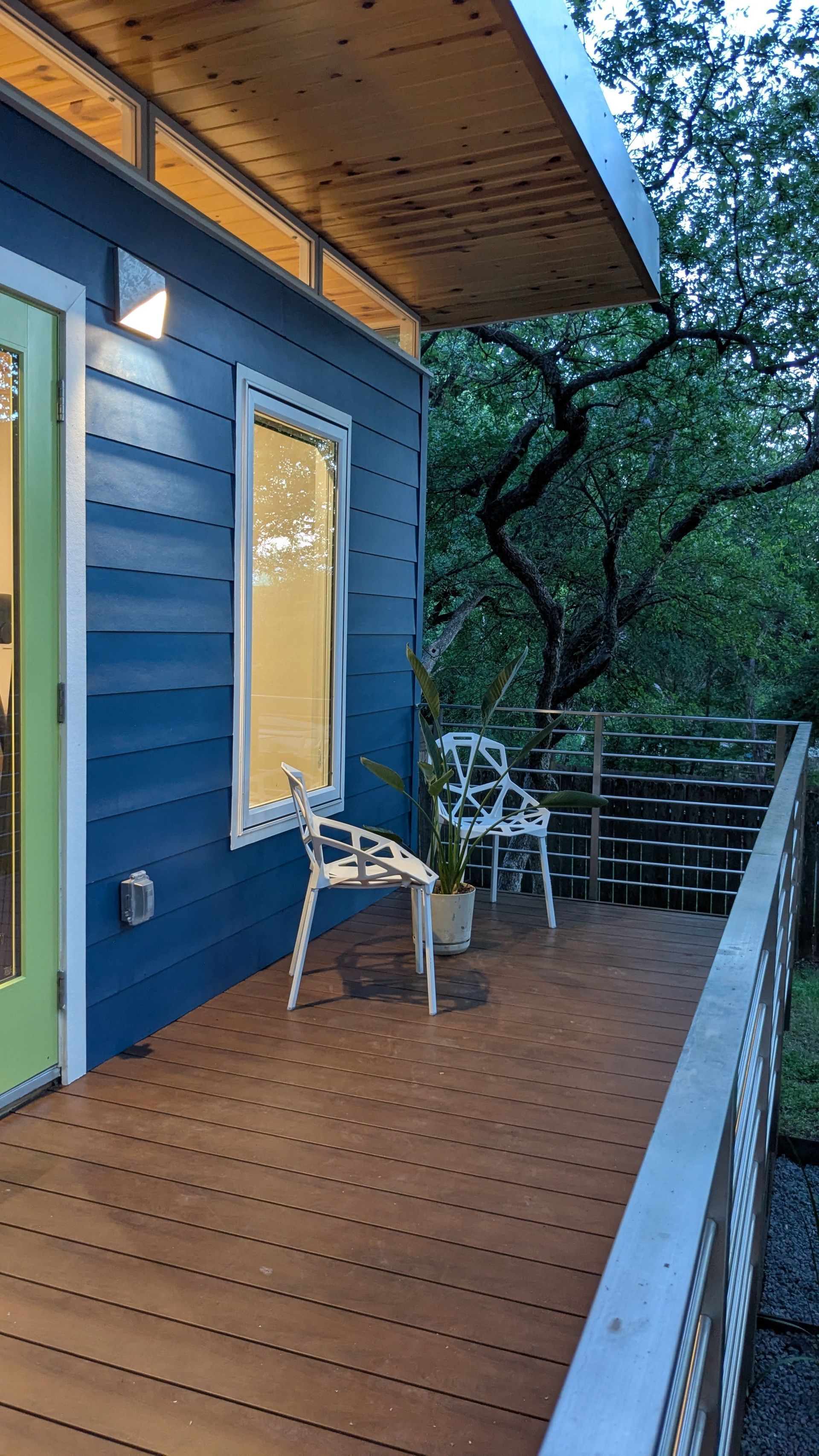 Blue house with wooden deck, white chairs, and a metal railing. Trees are in the background.