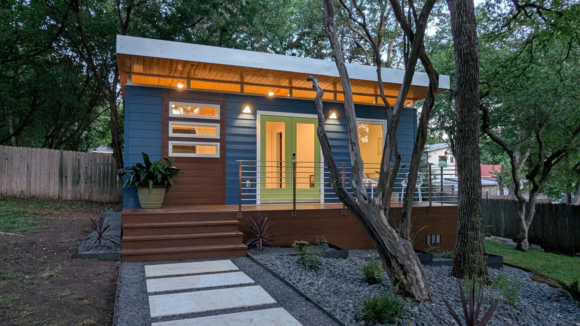 Blue tiny house with wood deck, accented with trees, gravel path, and foliage.