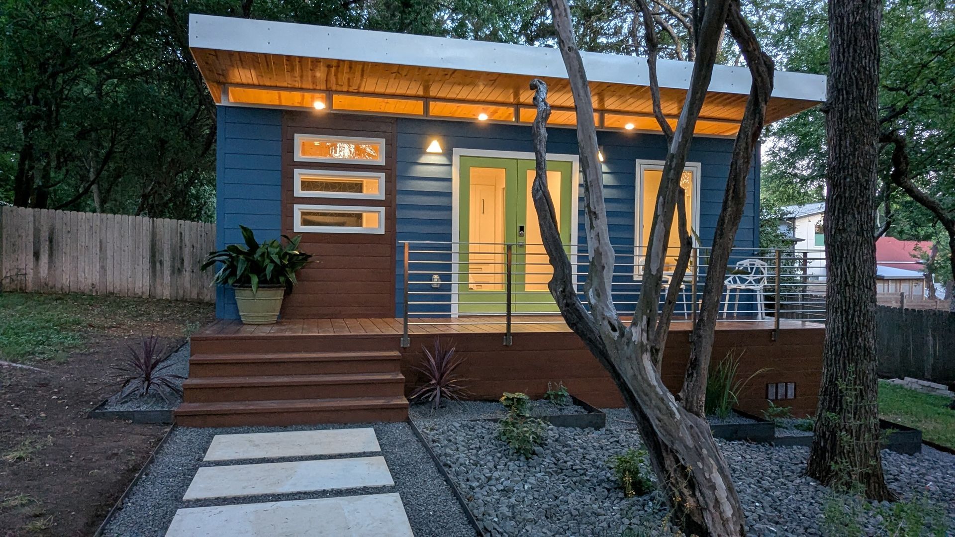 Blue modern small house with a white roof, wooden deck, and stepping stones leading to the entrance.