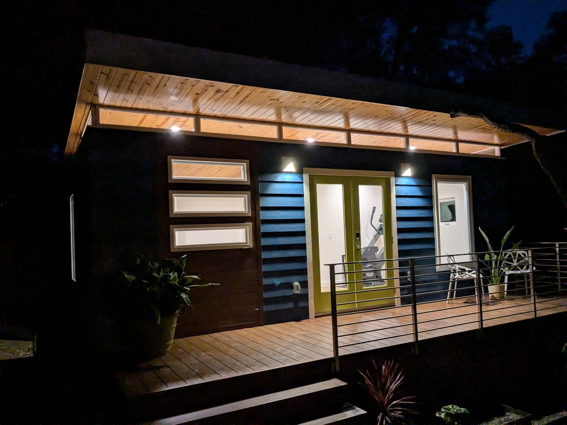 Blue house with wooden deck, at night. Illuminated awning, glass doors, and two sets of windows.