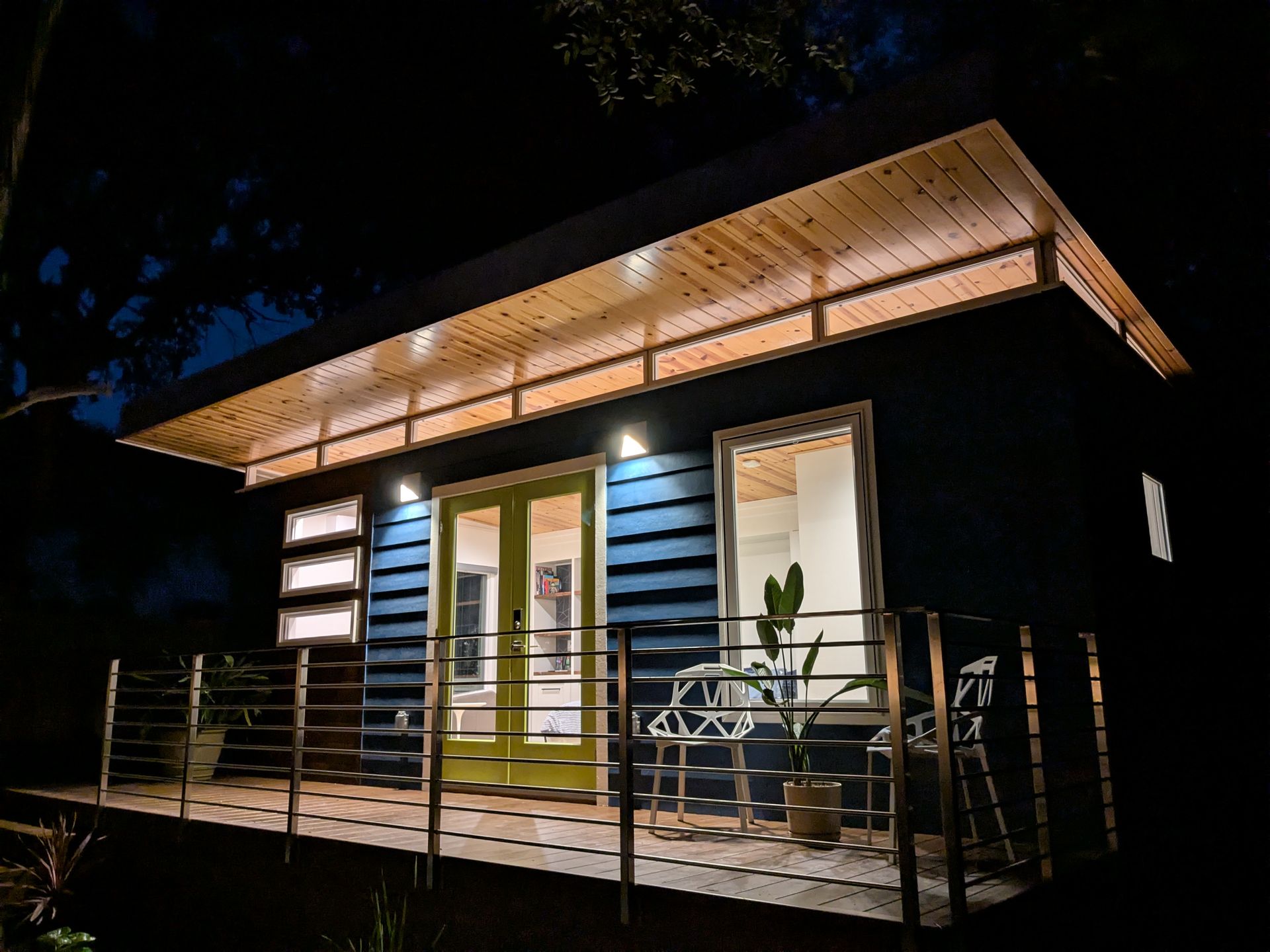 A modern, blue tiny home lit at night, with a wooden porch and a green door.