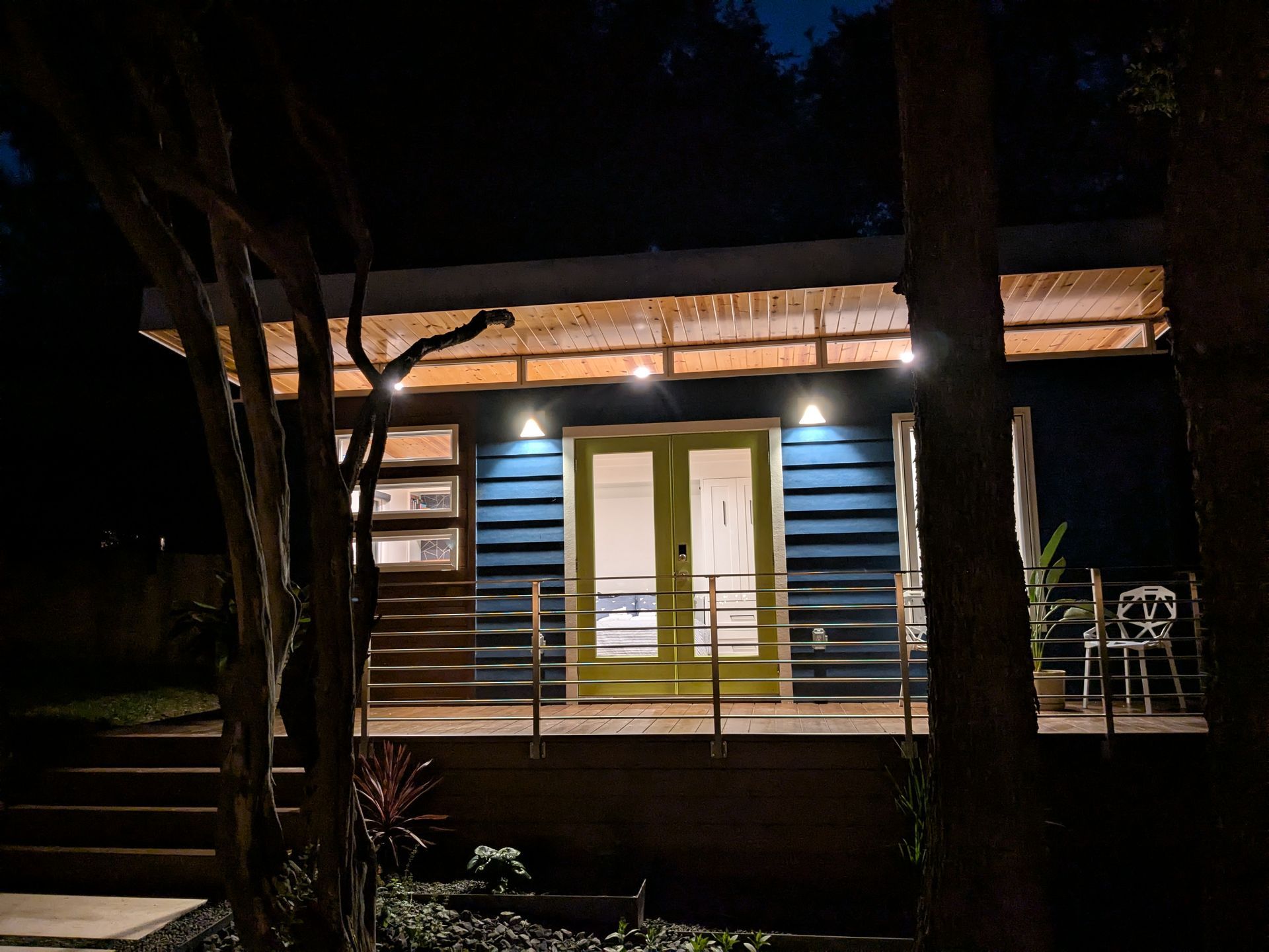 Night view of a blue-sided cabin with lit porch, green doors, and surrounding trees.